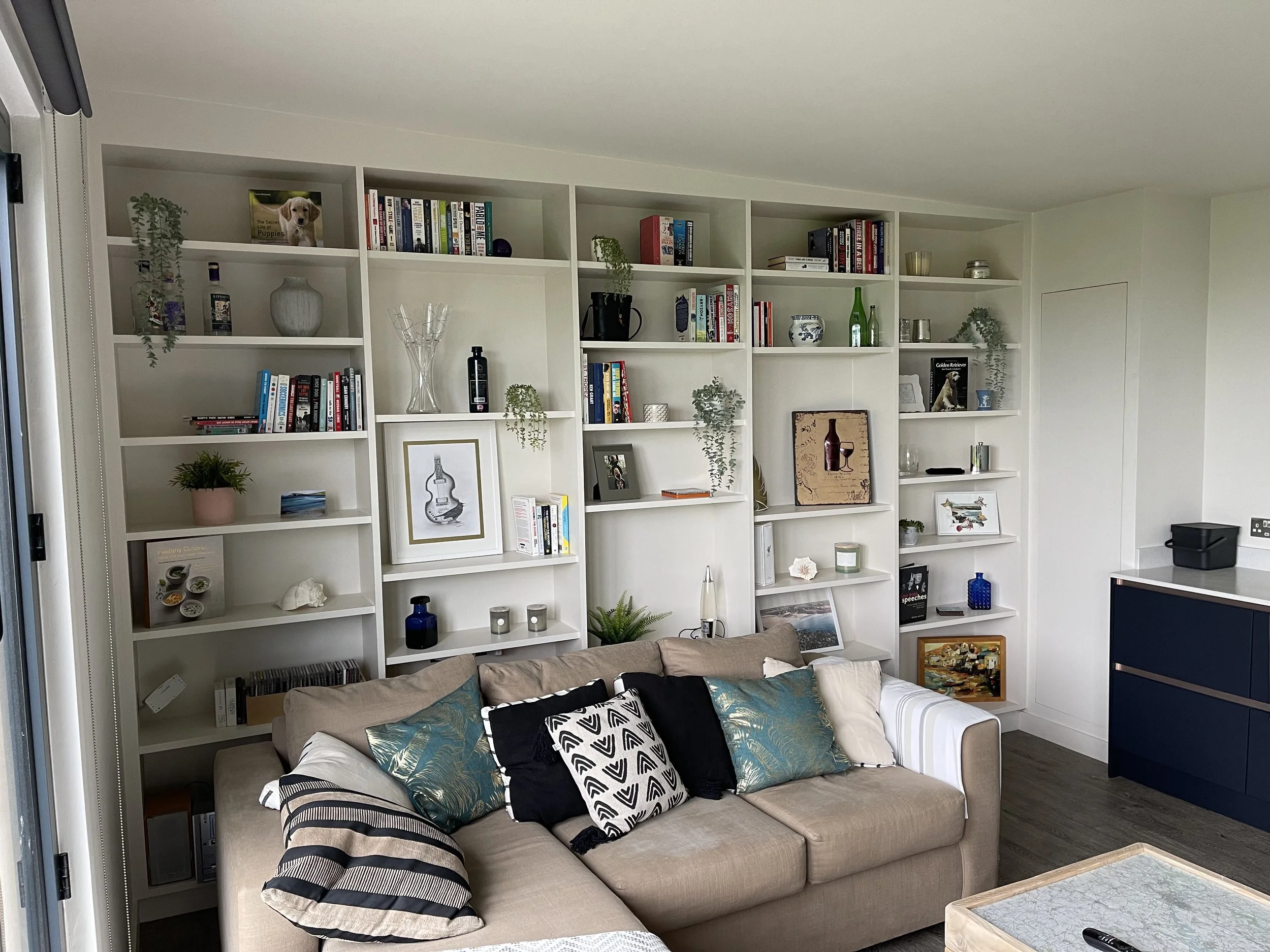 Living room with beige sofa adorned with patterned pillows, white bookshelves filled with books, decorative items, and plants, and a dark blue cabinet on the right side.