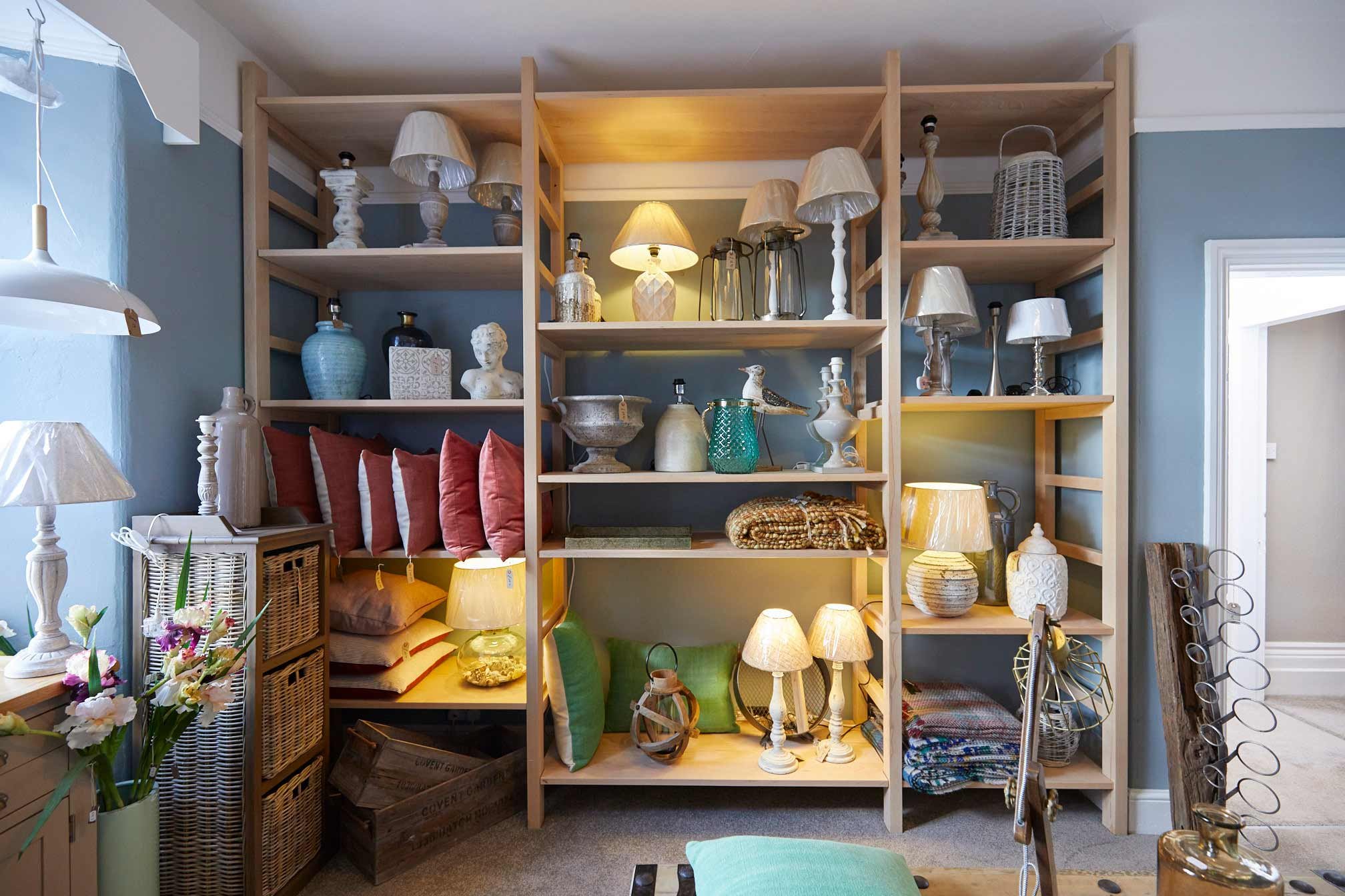 Open wooden shelves filled with lamps, decorative vases, sculptures, bowls, and textiles in a living room.