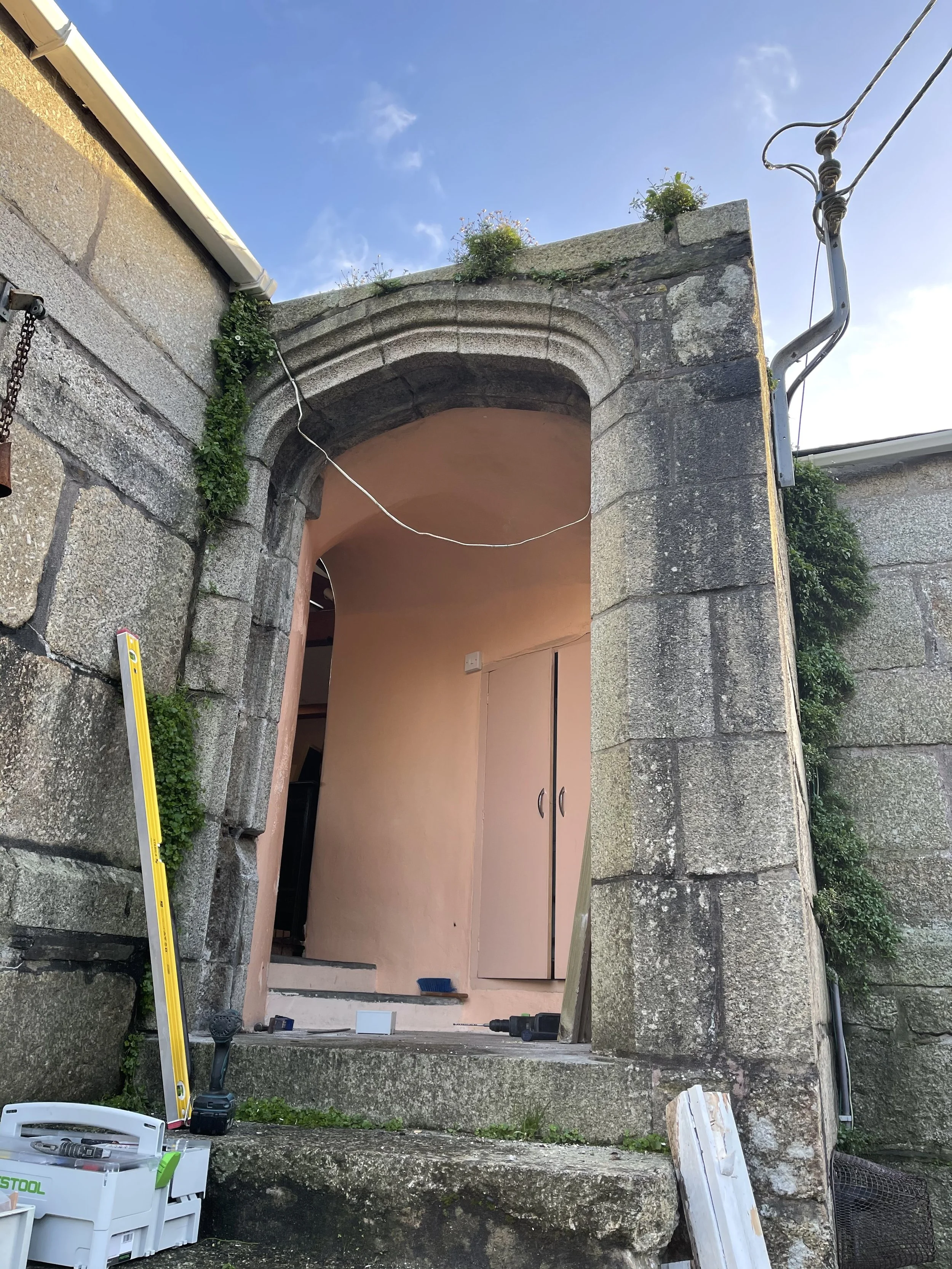 The image shows an outdoor stone archway entrance with tools and equipment nearby, and a small interior space with pinkish walls and closed cabinets inside.