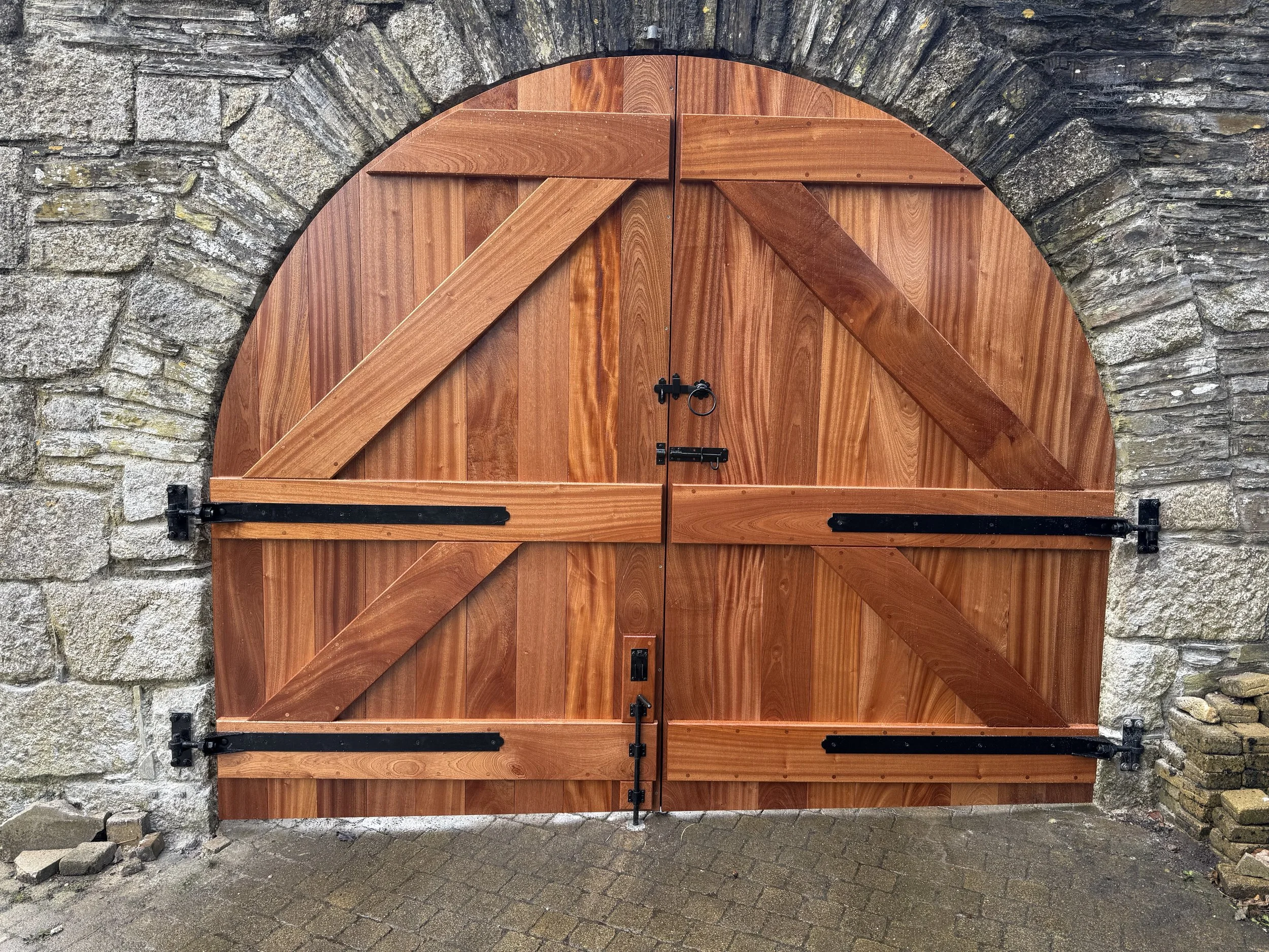 A large wooden gate with a rounded top, mounted on a stone wall with black metal hinges and latch, situated on a brick-paved surface.