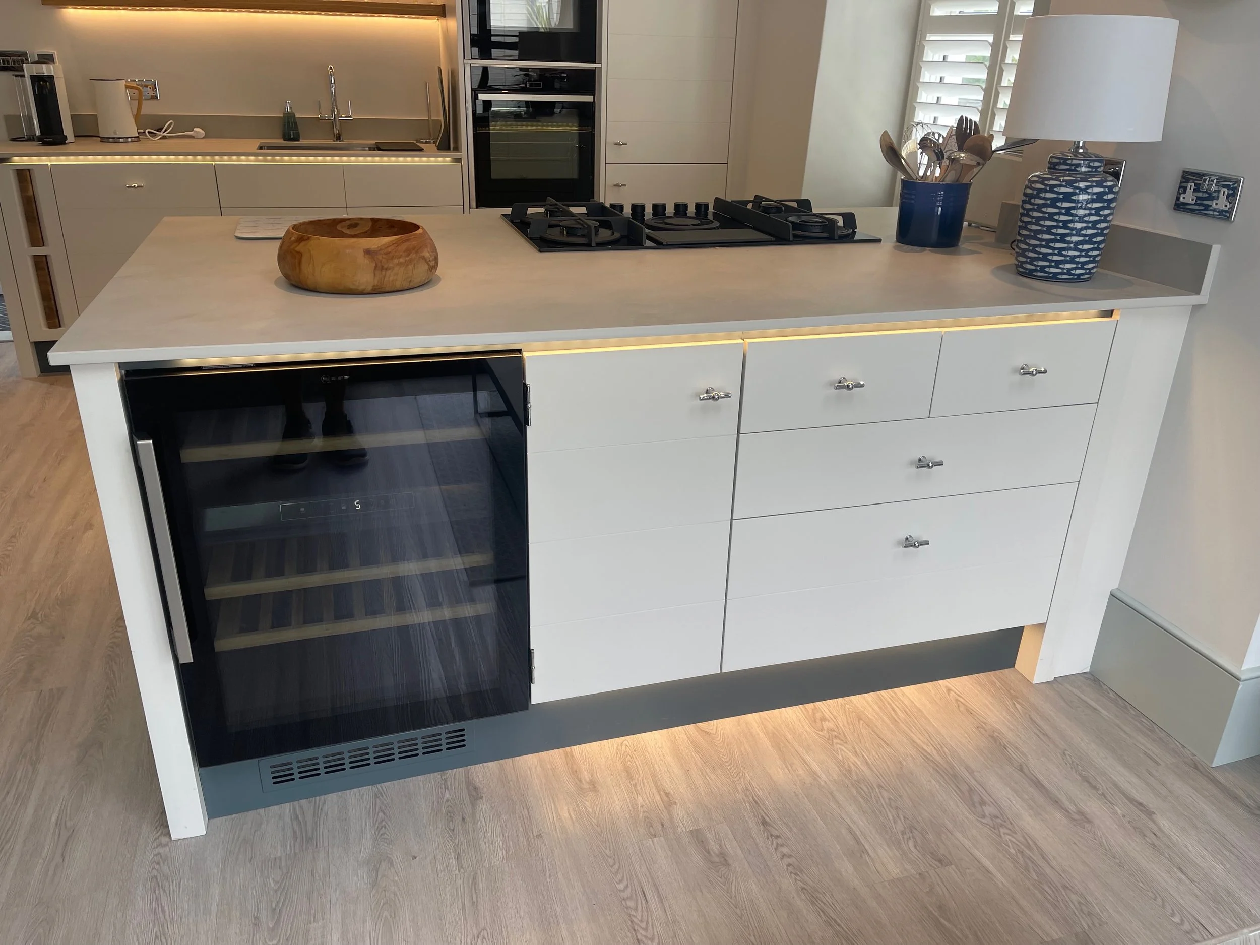Modern kitchen island with a built-in wine fridge, stovetop, and countertop accessories including a wooden bowl, a blue utensil holder, and a decorative vase with a lamp. Kitchen in the background with sink and appliances.