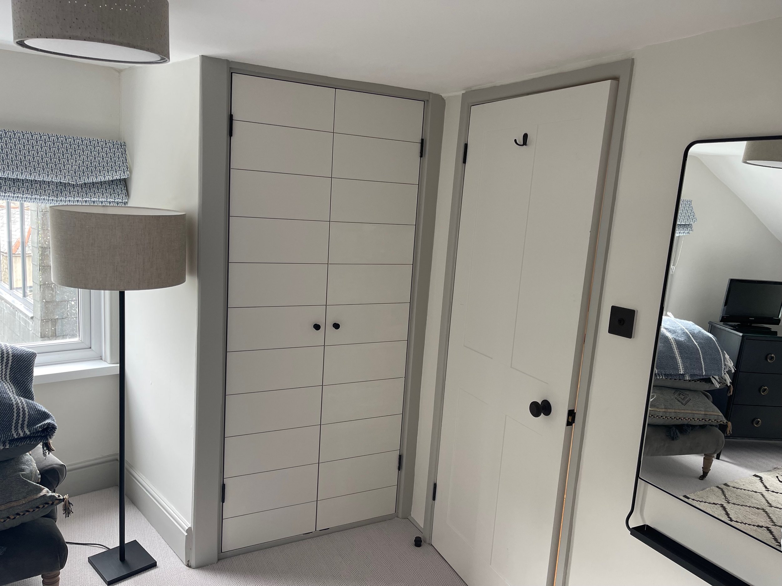 Corner of a room showing a window with a patterned Roman shade, a beige floor lamp, white double closet doors with black handles, a full-length mirror on the wall, and part of a bed with a black dresser and small TV in the background.