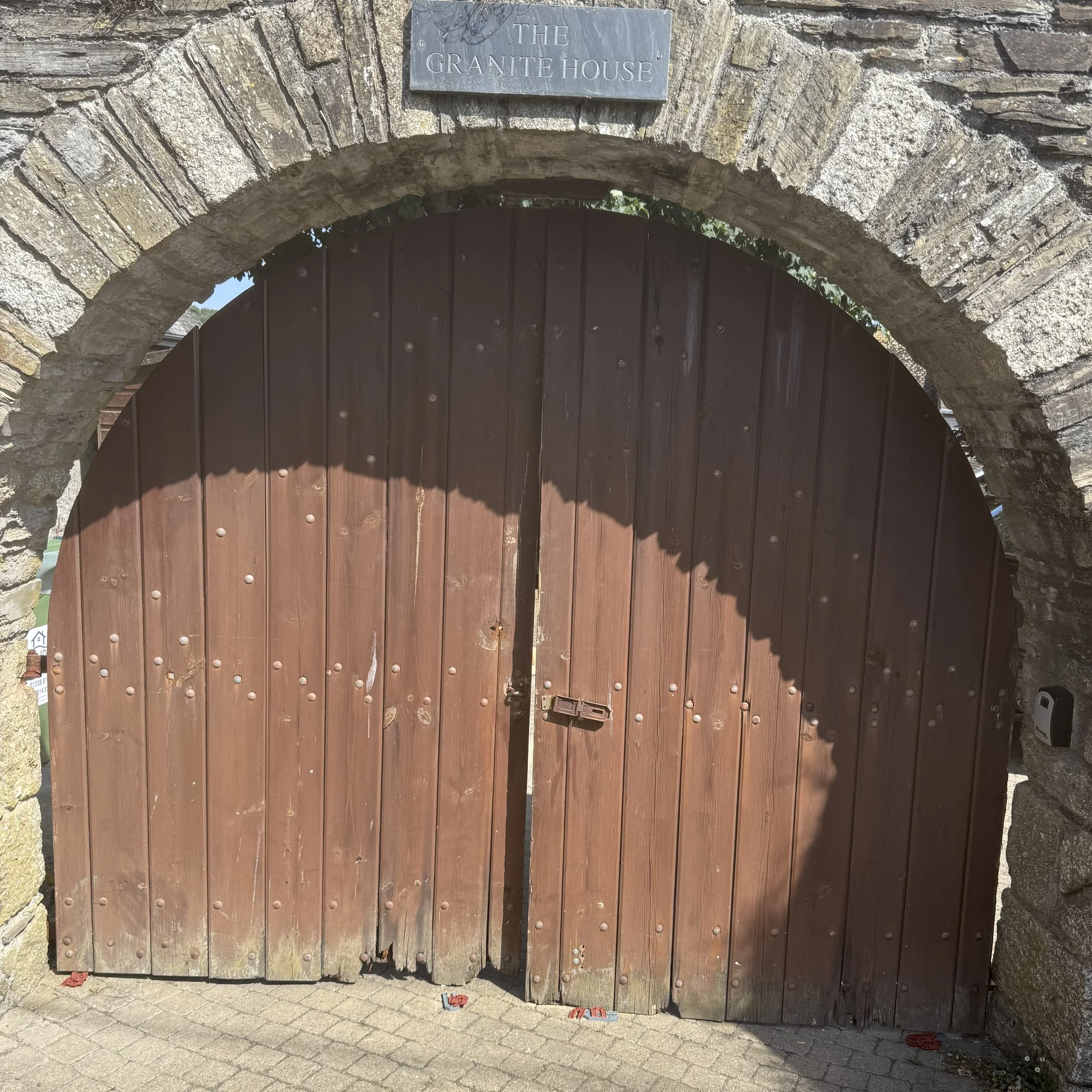 Wooden gate under a stone archway with a sign that reads 'The Granite House.' The gate has a latch and is set in a stone wall, with some shadows cast over it.