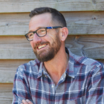 A man with a beard, glasses, and short dark hair wearing a plaid shirt, smiling, and sitting outdoors in front of a wooden background.