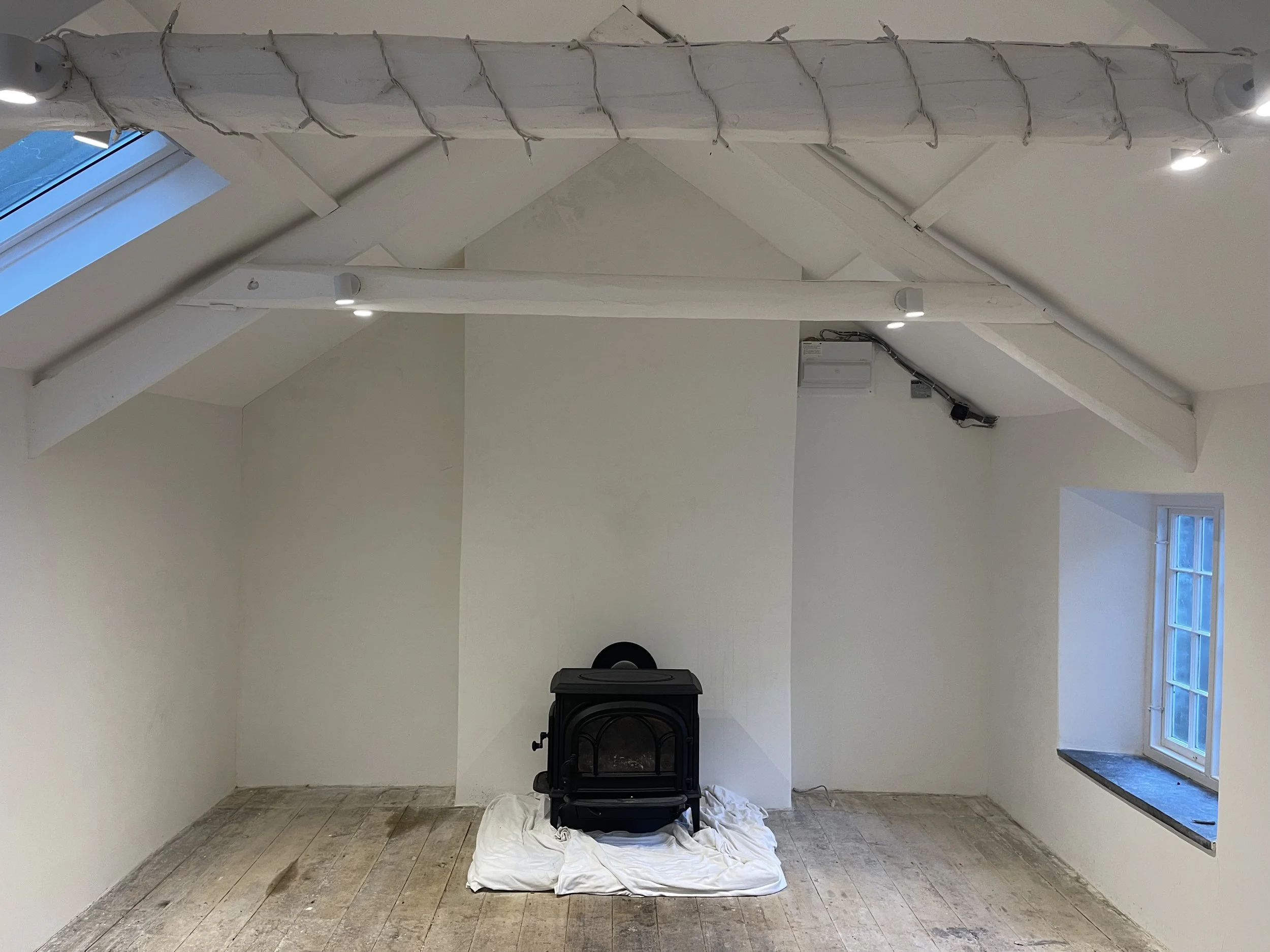 Empty attic room with white walls and wooden floor, a black stove on a white cloth, a skylight window on the left, and a small window on the right wall. Exposed ceiling beams and string lights are present.