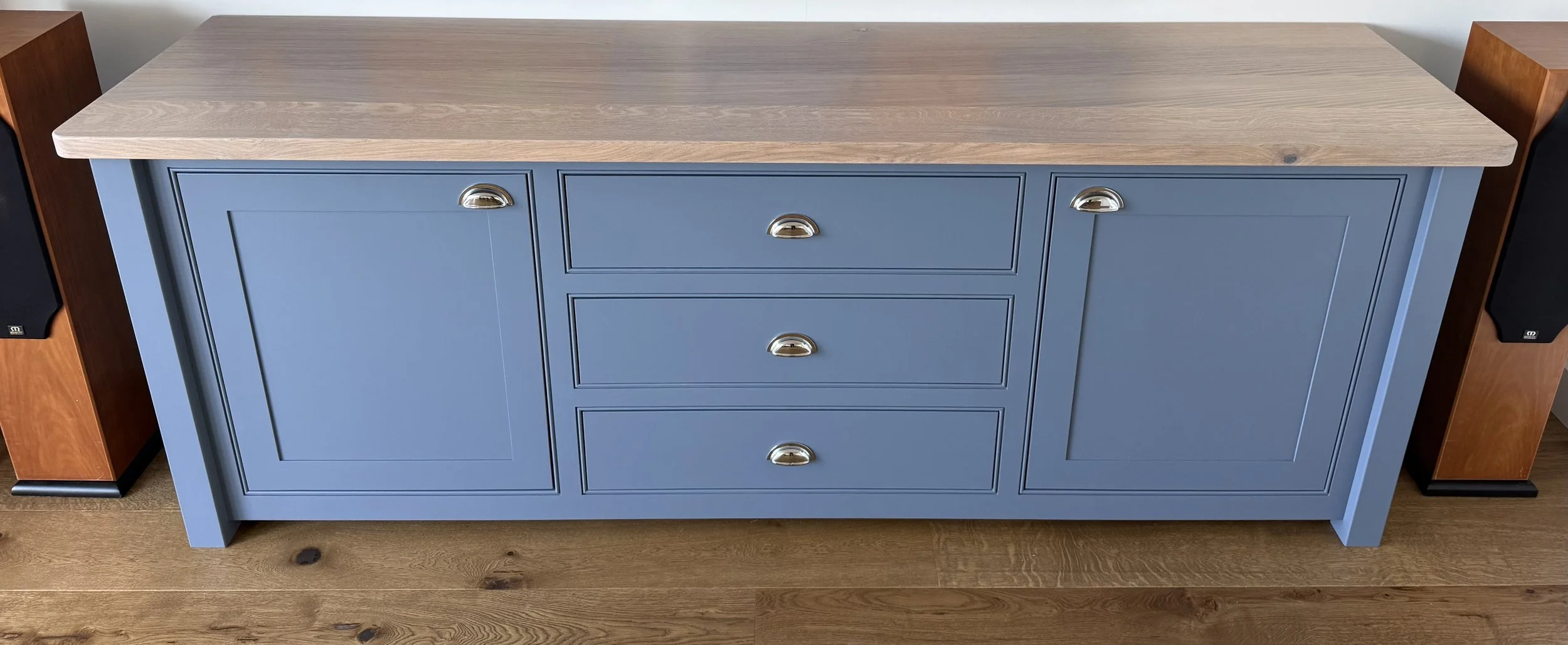 Blue wooden sideboard with a natural wood top and chrome handles, flanked by wooden speaker stands on a wooden floor.