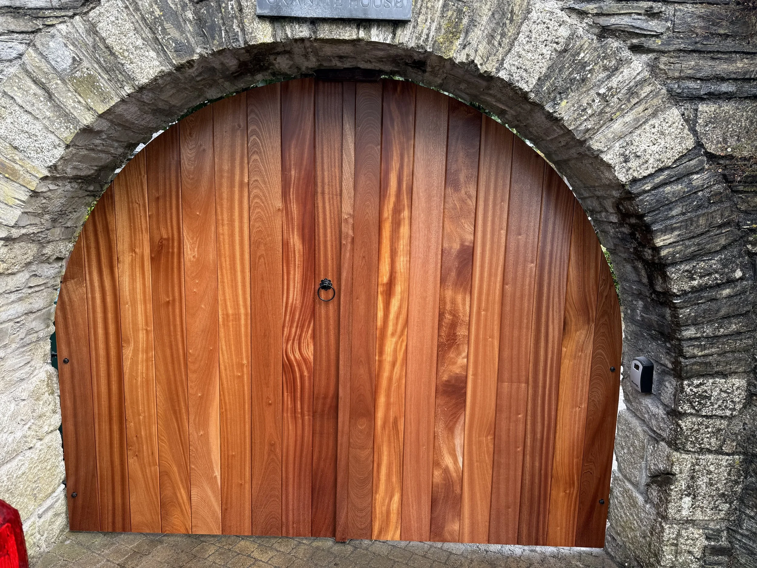 A wooden arched gate with vertical planks, set in a stone brick archway. There is a black ring handle and a remote control device on the stone wall to the right.