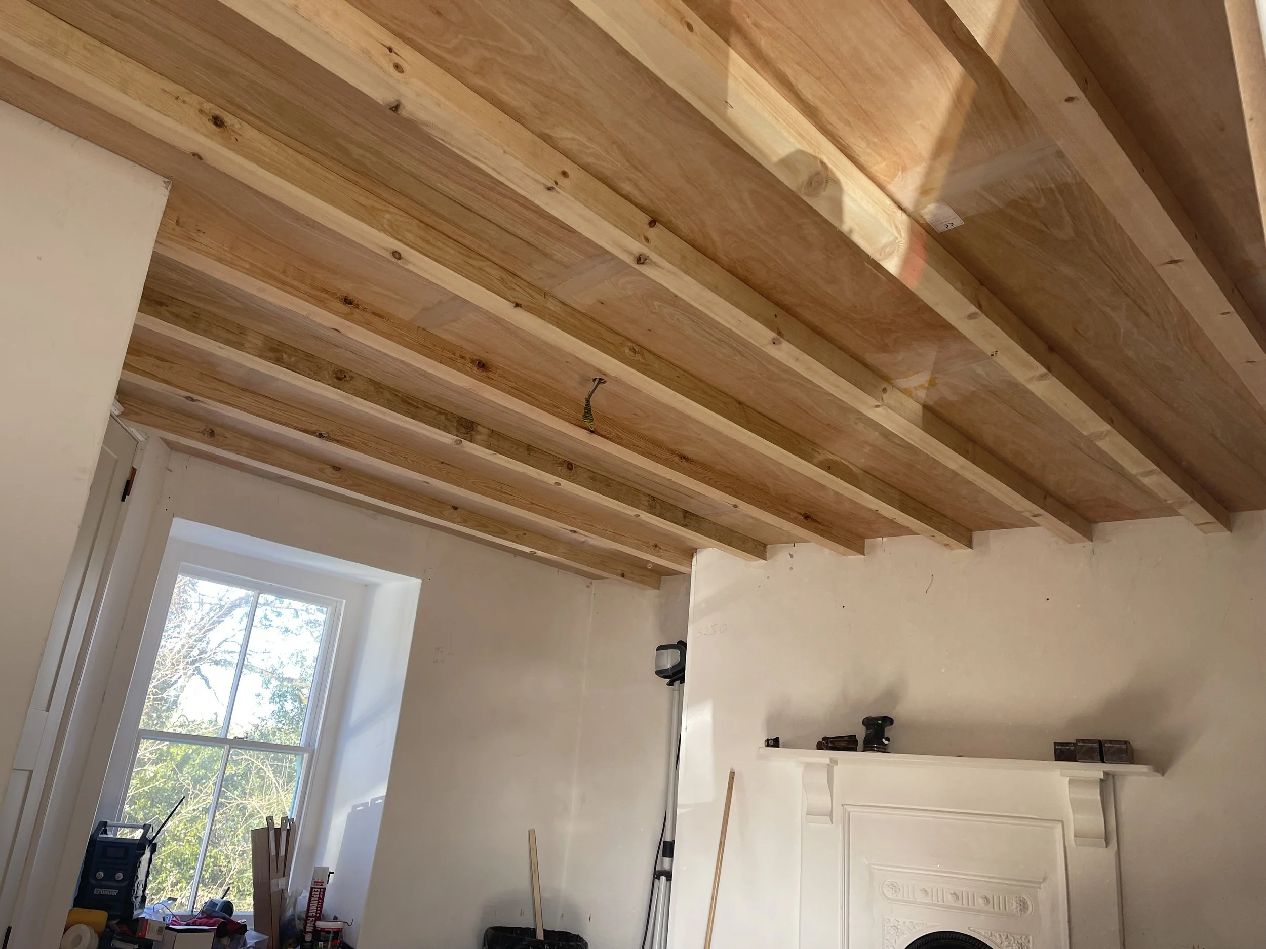Interior of a room under renovation with a wood-paneled ceiling, a window showing trees outside, and construction tools and materials on the windowsill and fireplace mantel.