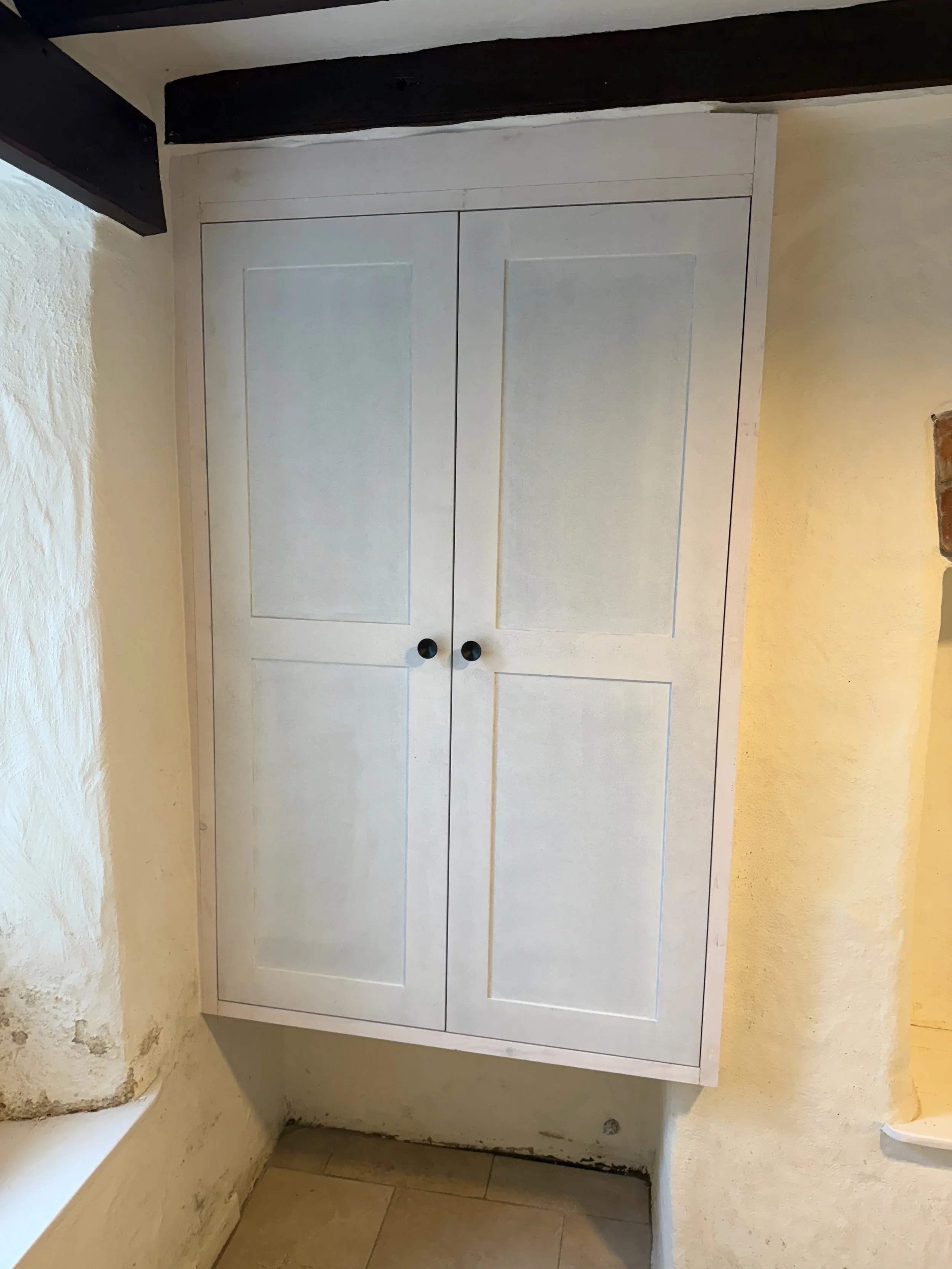 White cabinet with two doors and black round knobs in a rustic room with textured walls and wooden beams.
