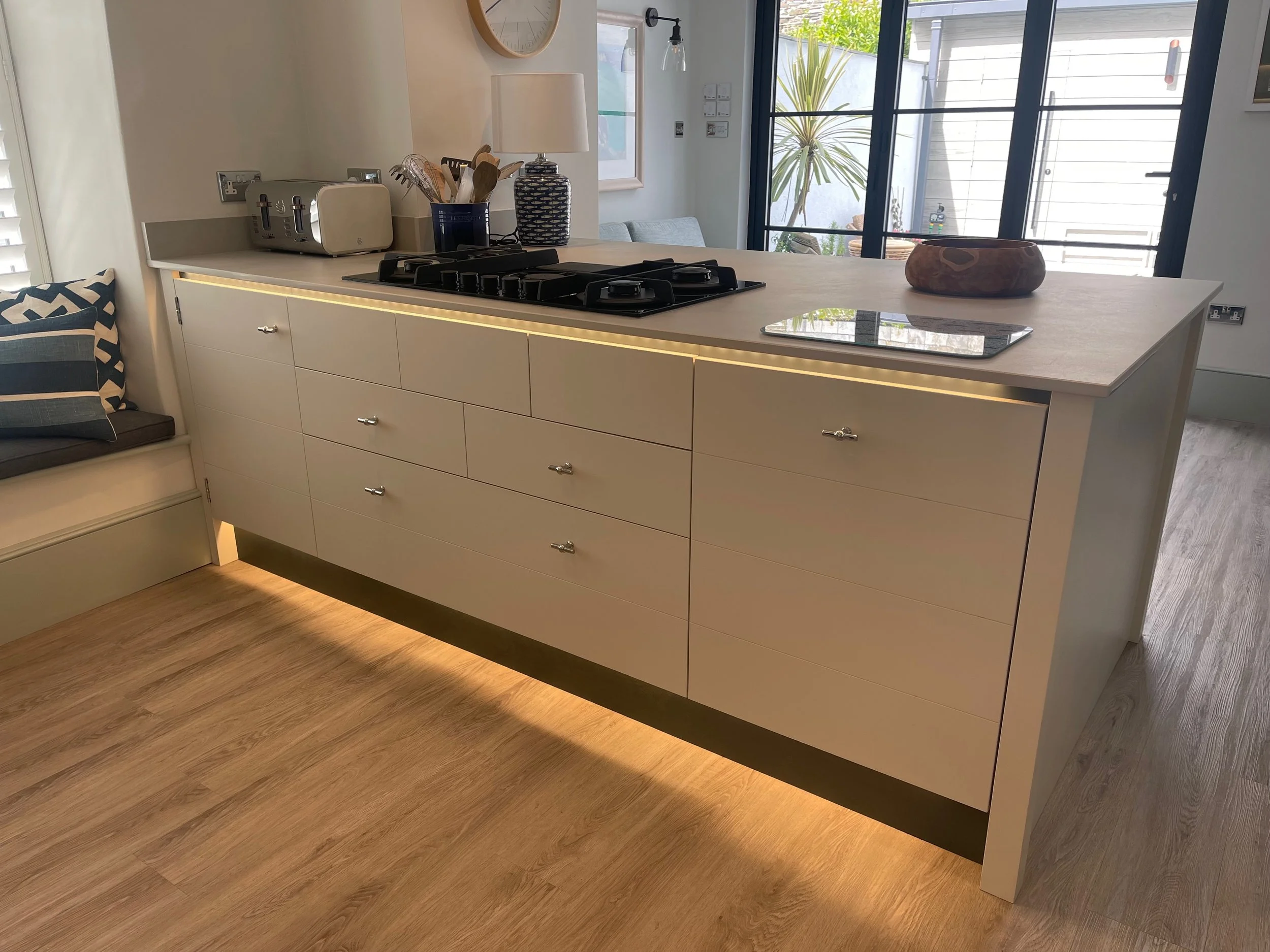 Modern kitchen island with drawers and a wooden countertop, with some kitchen appliances and utensils on top, a windowed door in the background, and a bench with cushions to the side.