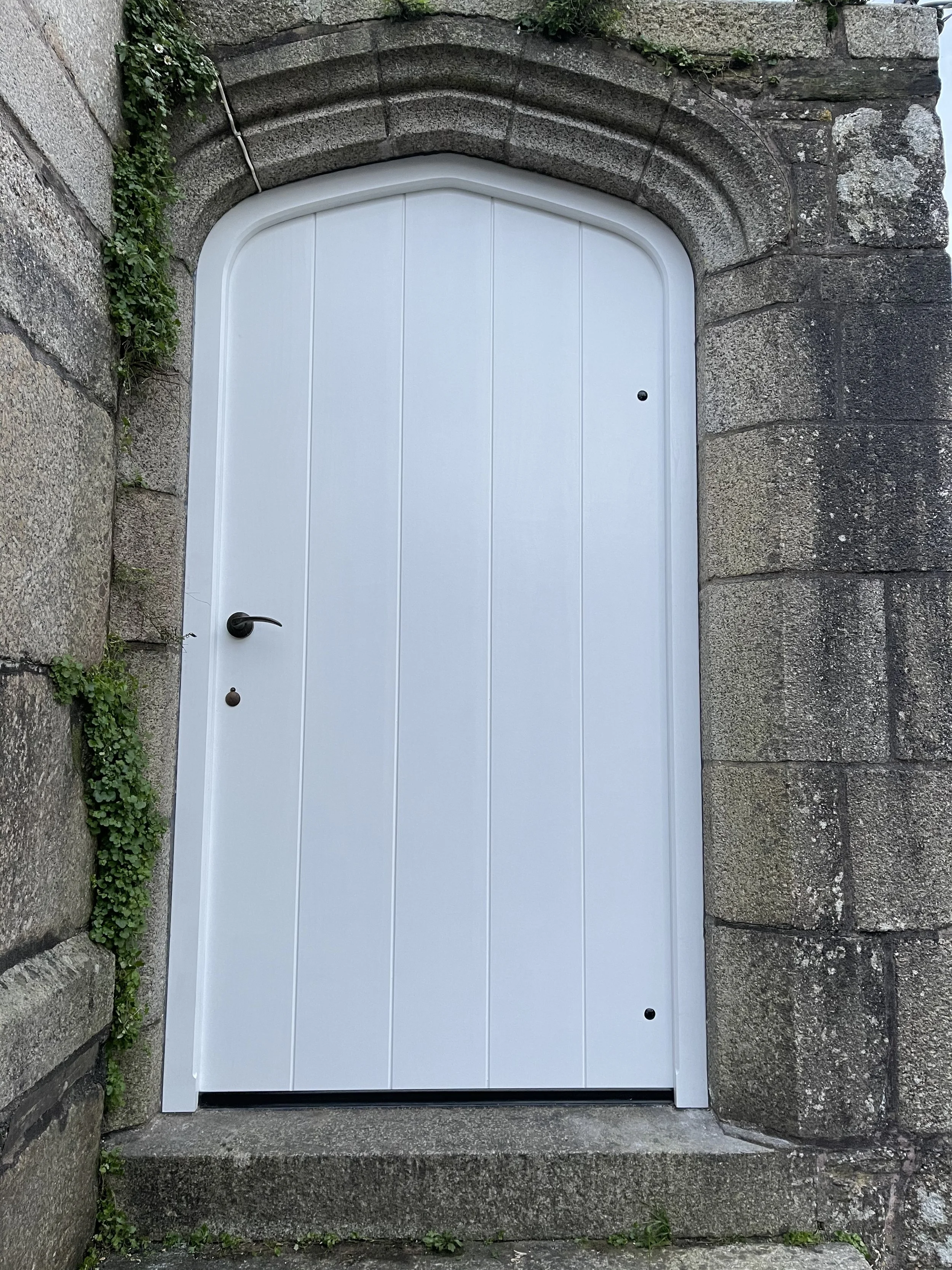 A white door set within a stone archway, surrounded by stone walls, with a black handle and a small peephole.