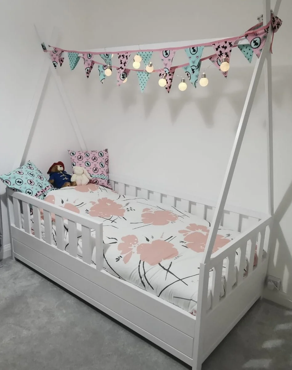 Children's bed with pink and blue triangular bunting, decorative string lights, and plush toys, in a white children's room.