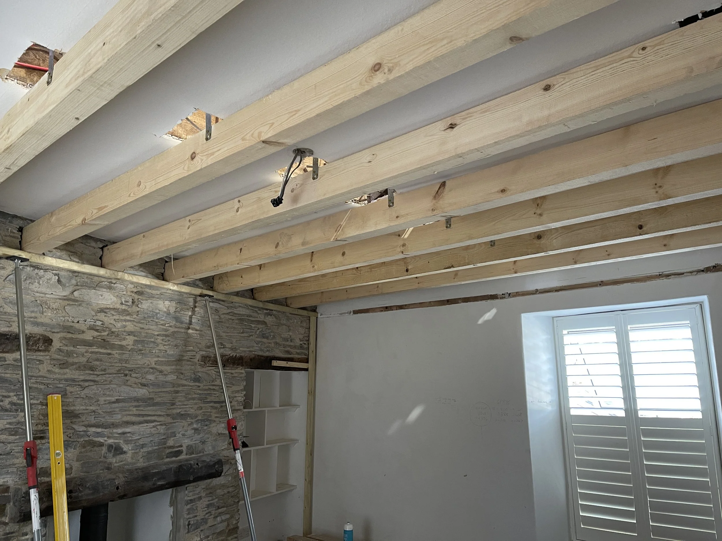 Interior of a room under renovation with exposed wooden ceiling beams, a stone wall, a window with shutters, and construction tools standing in the room.