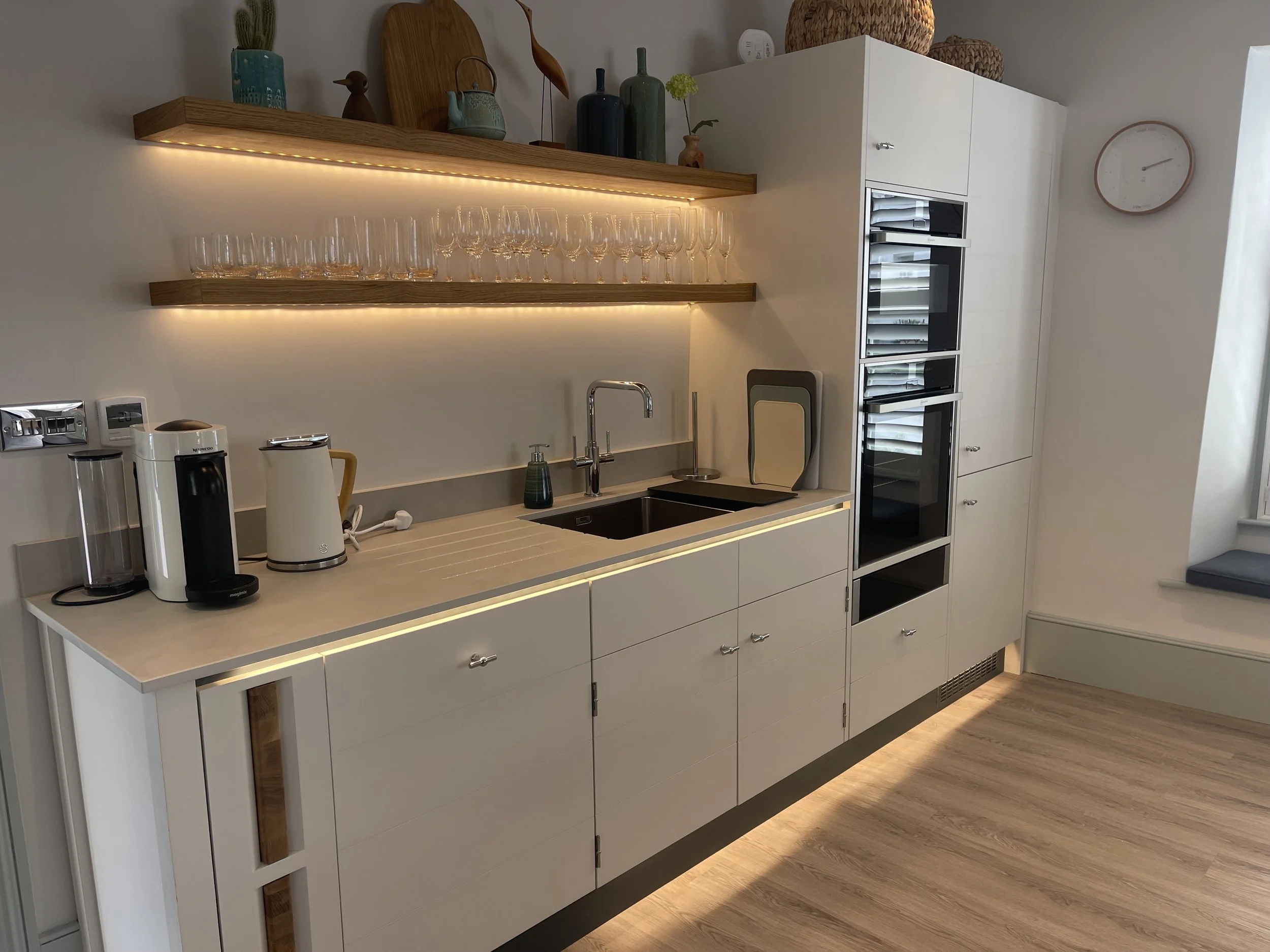 Modern kitchen with white cabinets, wooden shelves, glassware, and built-in appliances. Decor includes vases and woven baskets, with LED lighting under the shelves providing ambient light.