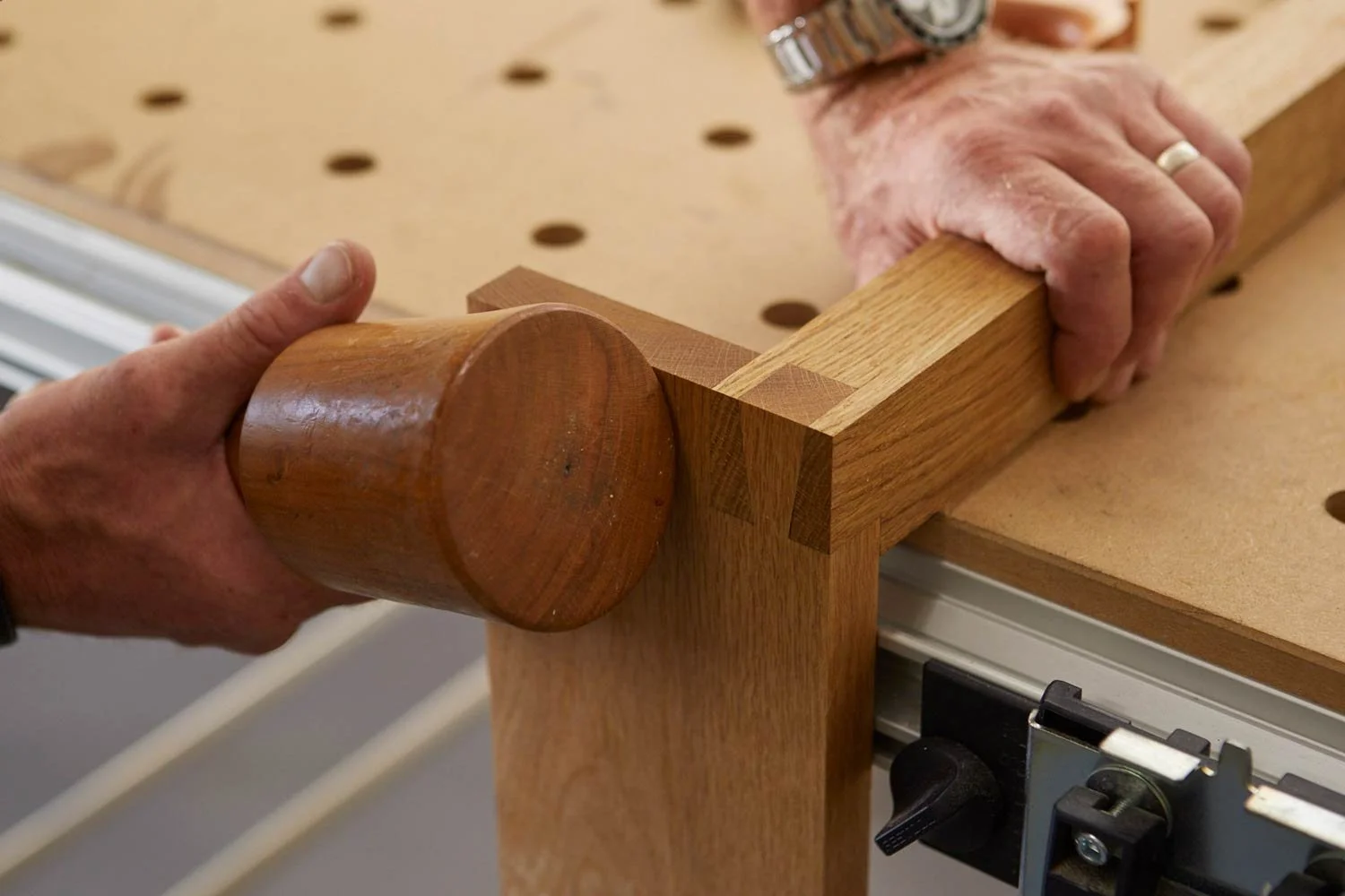 A person using a mallet to assemble a wooden piece on a workbench, with a woodworking vise and holes in the work surface visible.
