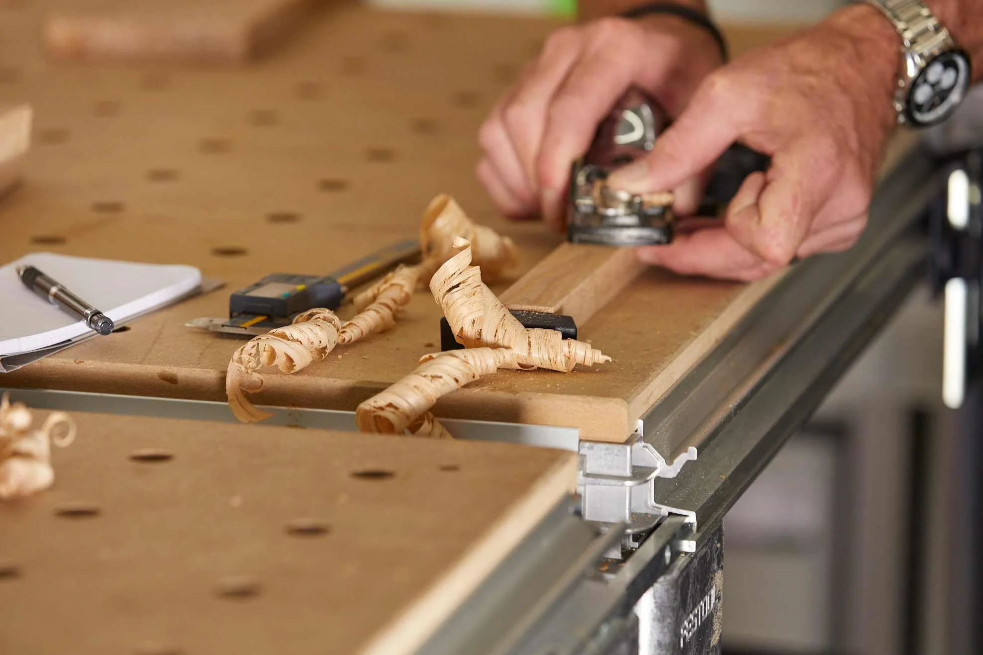 Someone using a hand planer on a piece of wood, with wood shavings on a woodworking workbench.
