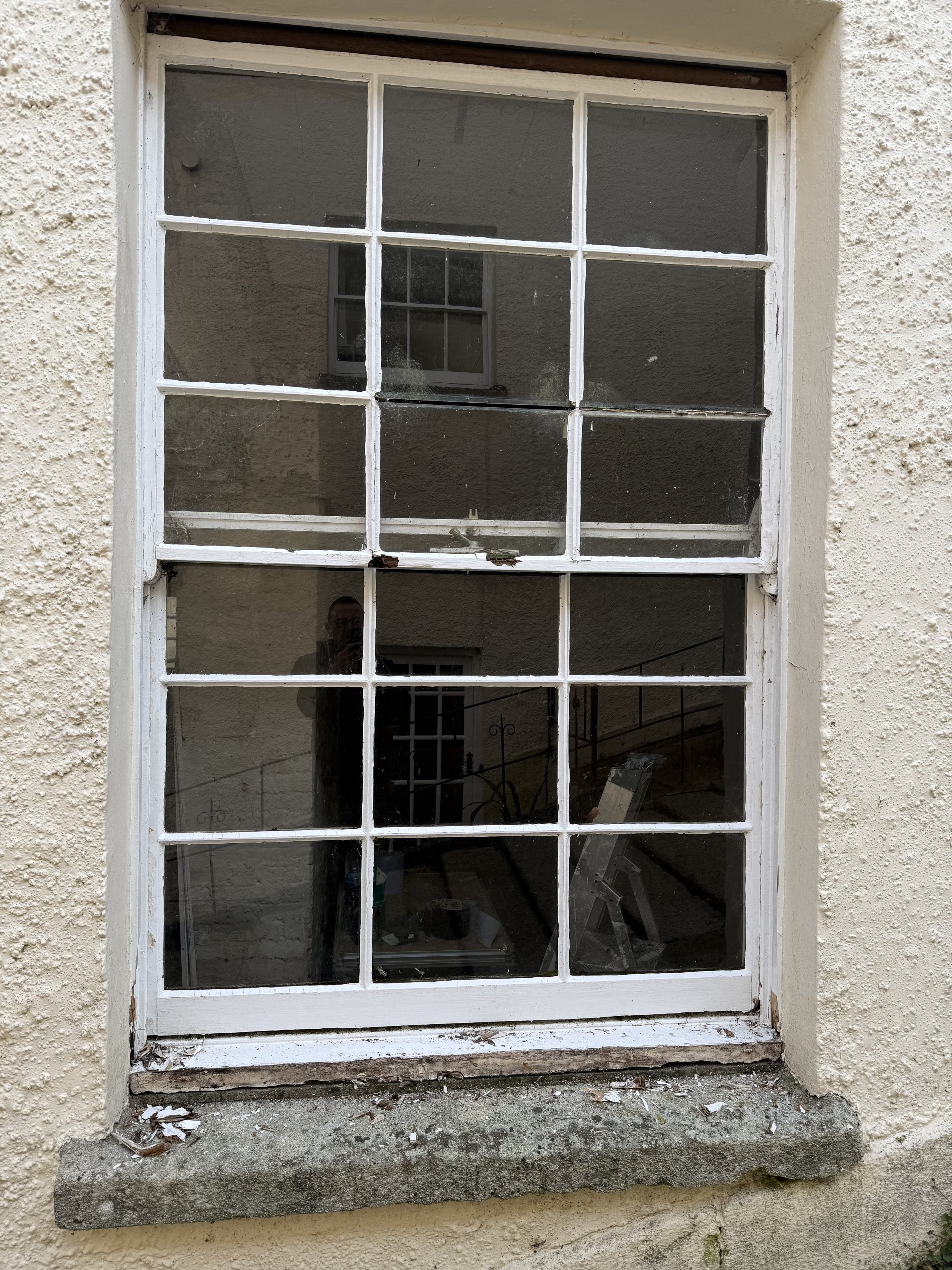 The image shows a window with a white wooden frame, having two horizontal panes and three vertical panes, set in a yellowish, textured stucco wall. The window appears old with some paint chipping and minor damage at the bottom frame. The reflection i