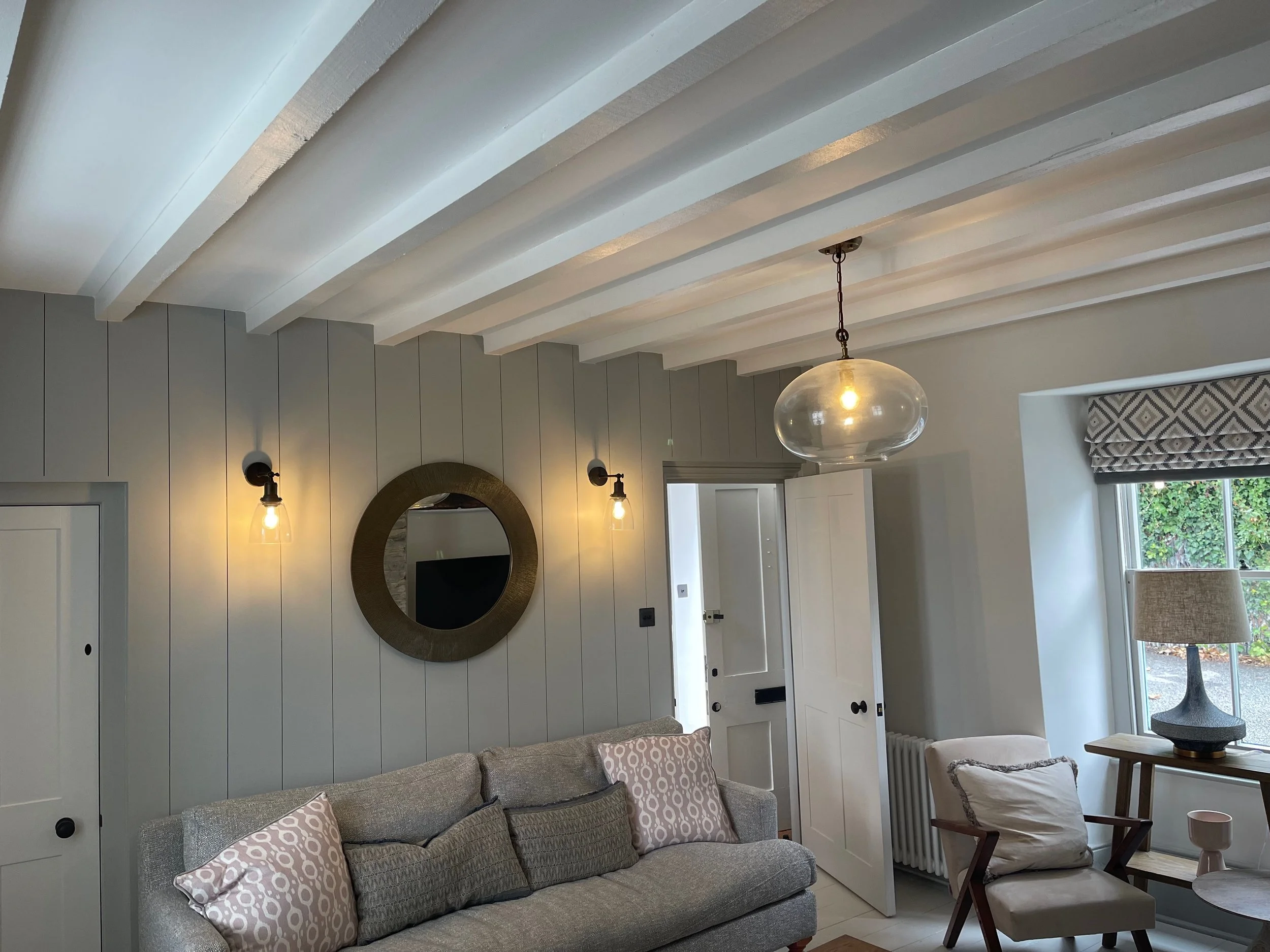 Living room with gray sofa, patterned throw pillows, wooden side chair, round window, wall mirror, side table with lamp, and glass pendant light hanging from white ceiling beams.