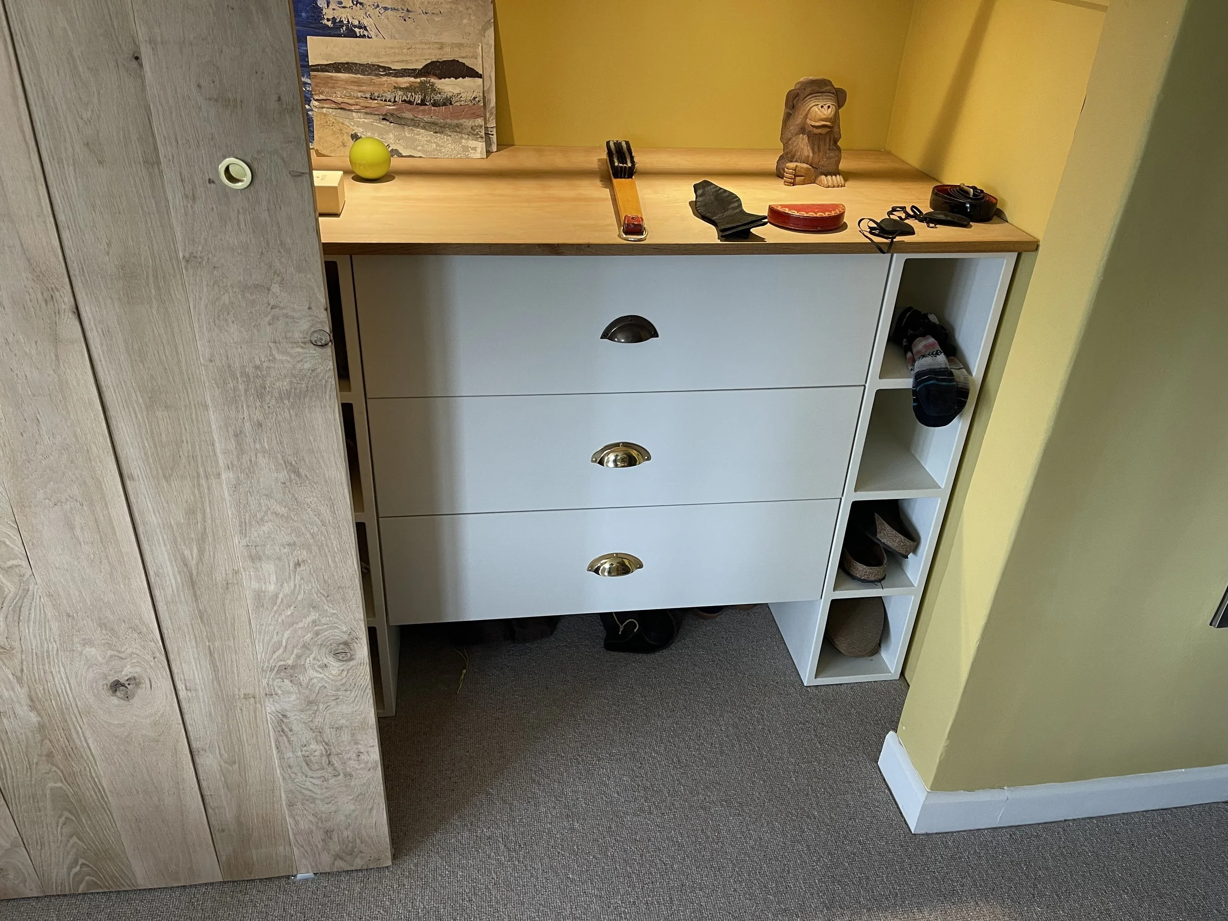 A white multifunctional storage unit with drawers and open cubbies against a yellow wall, topped with various small items including a sculpture, remote, and papers; a beige and wooden cabinet to the left.