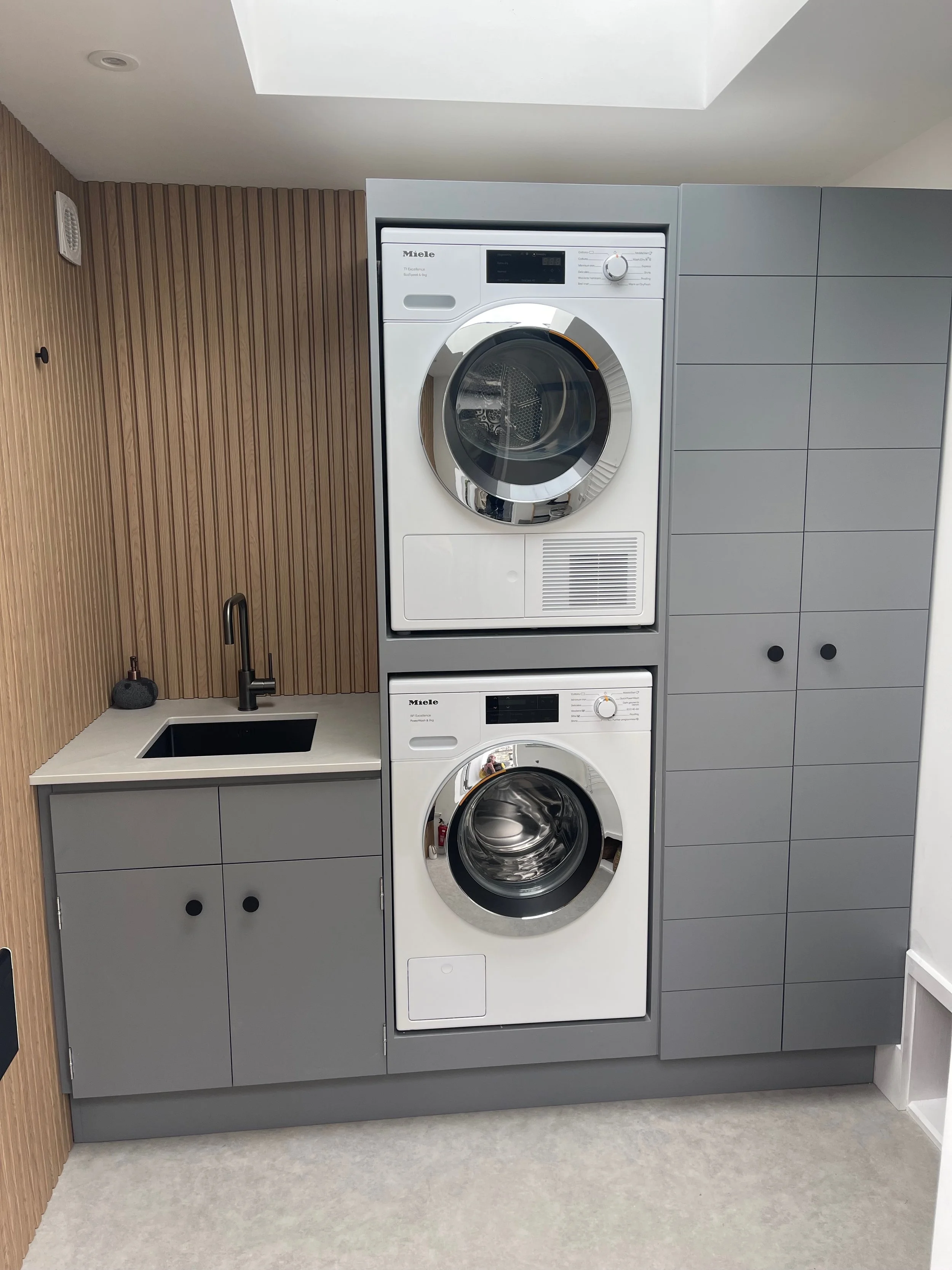 A laundry room with stacked front-loading washer and dryer, a small sink with a black faucet, wooden wall paneling, and gray cabinets.