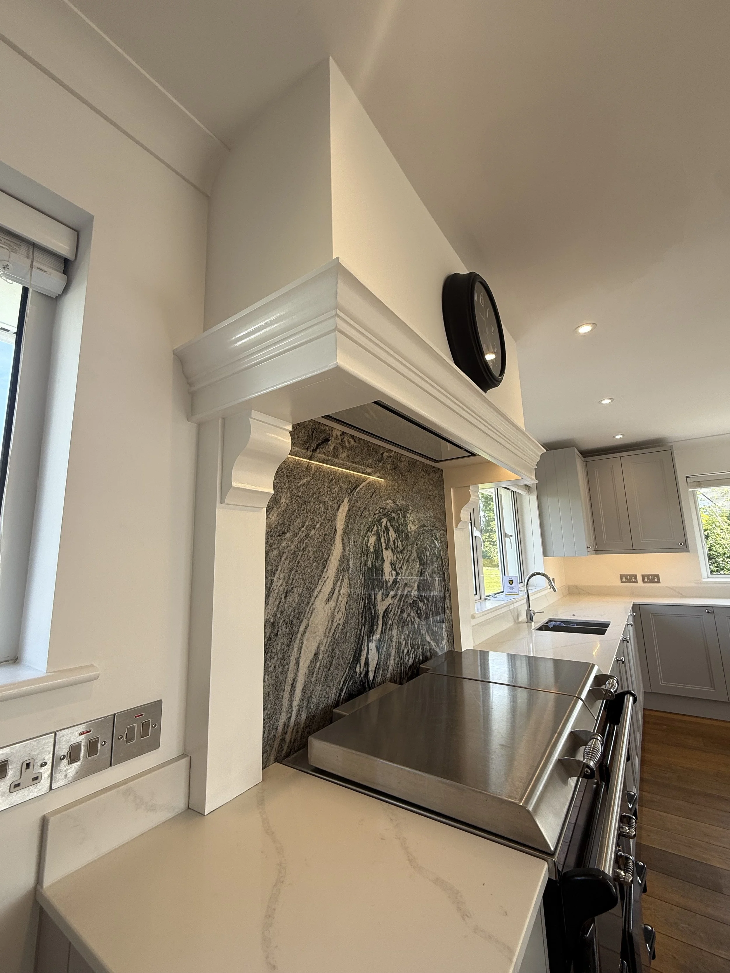 Modern kitchen with cream-colored countertops, white cabinets, and a dark granite backsplash with a marbled pattern. Includes a stainless steel oven, a window above the sink, and a black wall clock.