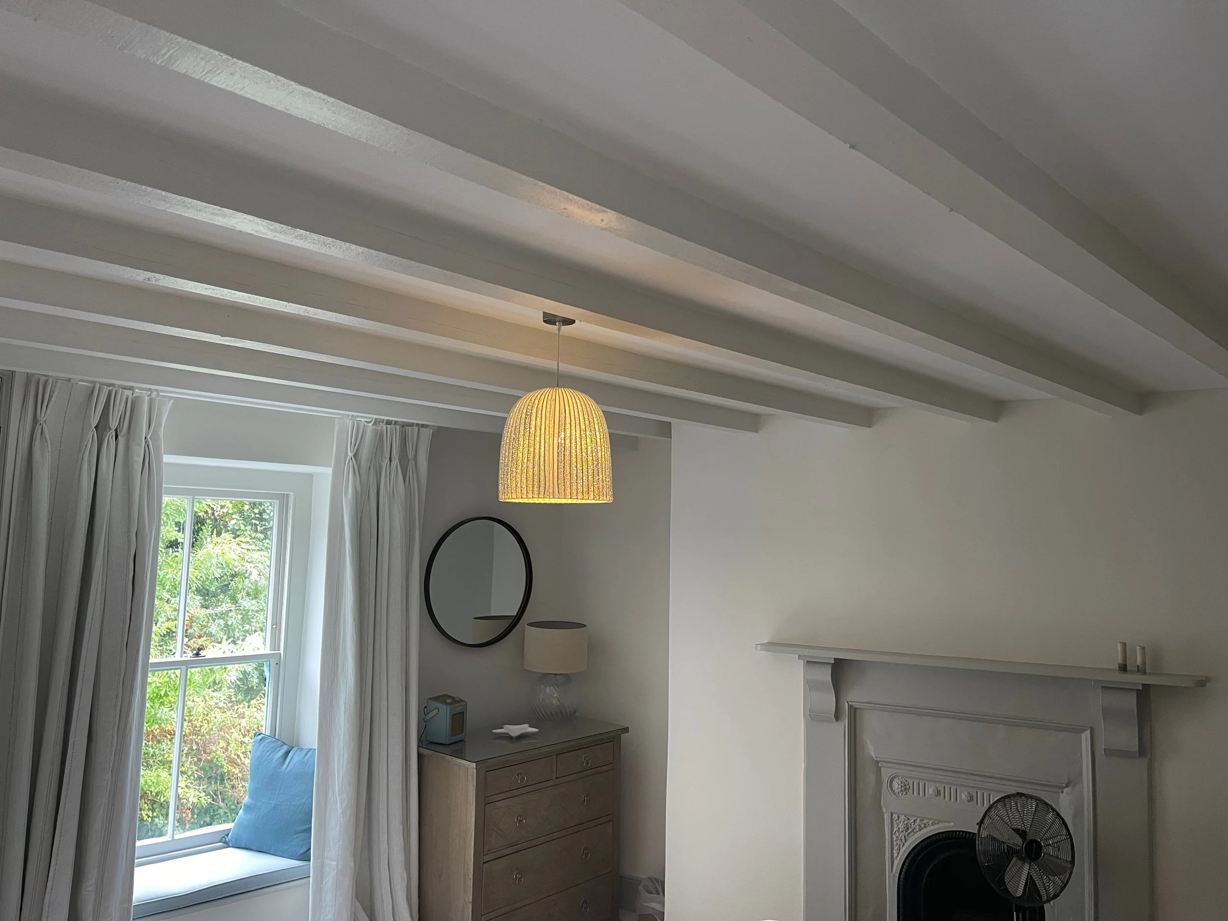 Living room with white ceiling beams, a hanging yellow lamp, a window with white curtains, a dresser with a lamp, a mirror, and a small fan in front of a fireplace.