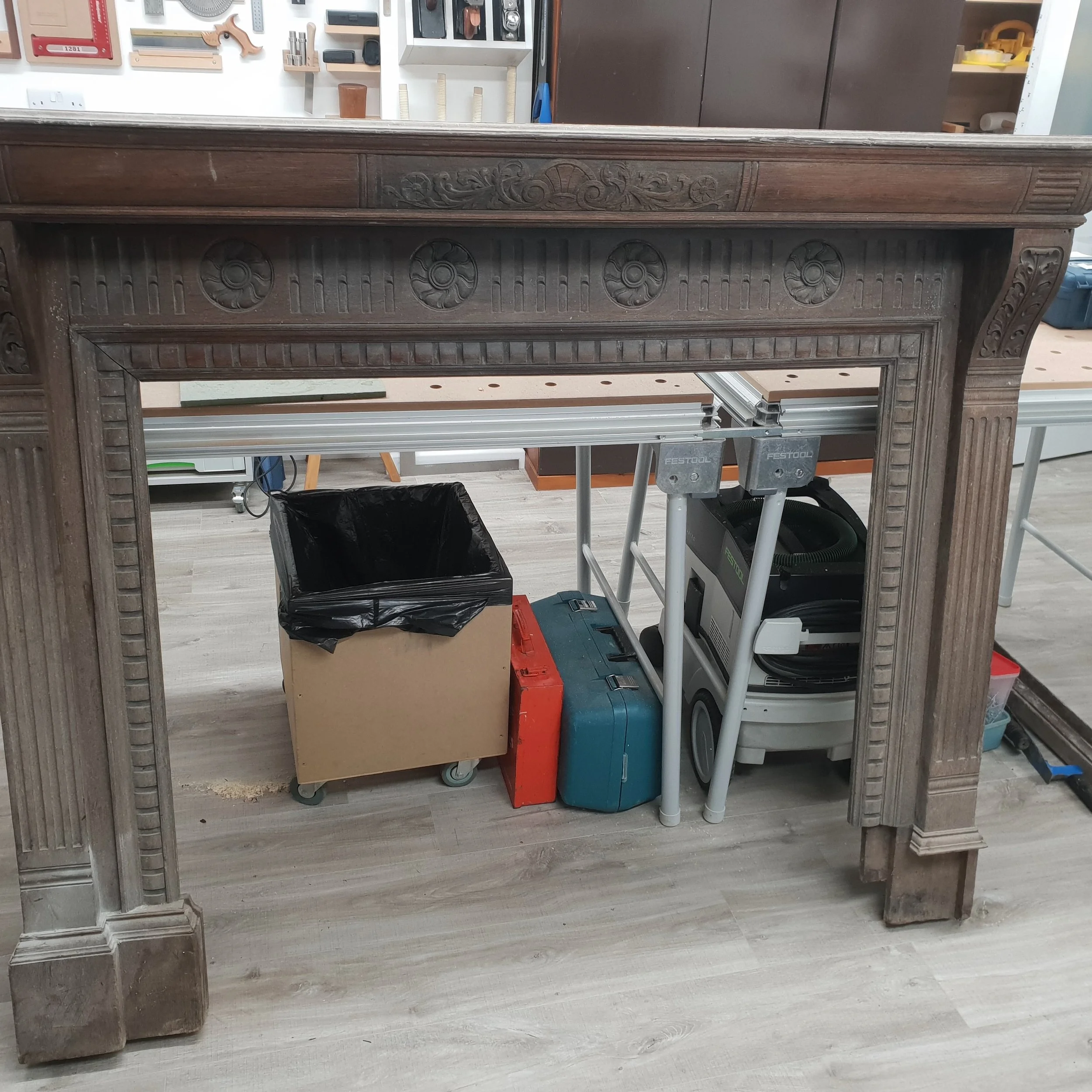 A wooden fireplace mantel with intricate carved details and floral designs, situated in a workshop with tools and equipment in the background.
