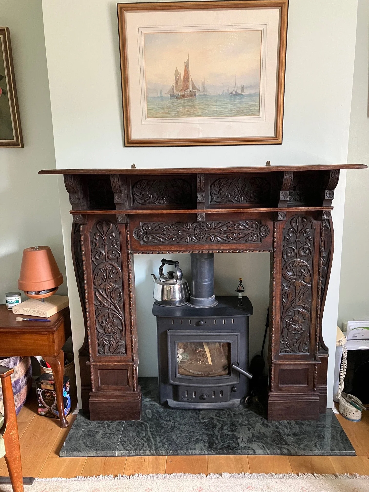 A wooden mantel with intricate carvings surrounds a black wood stove. On top of the mantel, there is a metal kettle and a chimney pipe. Above the mantel, a framed watercolor painting of sailboats on water hangs on the wall.