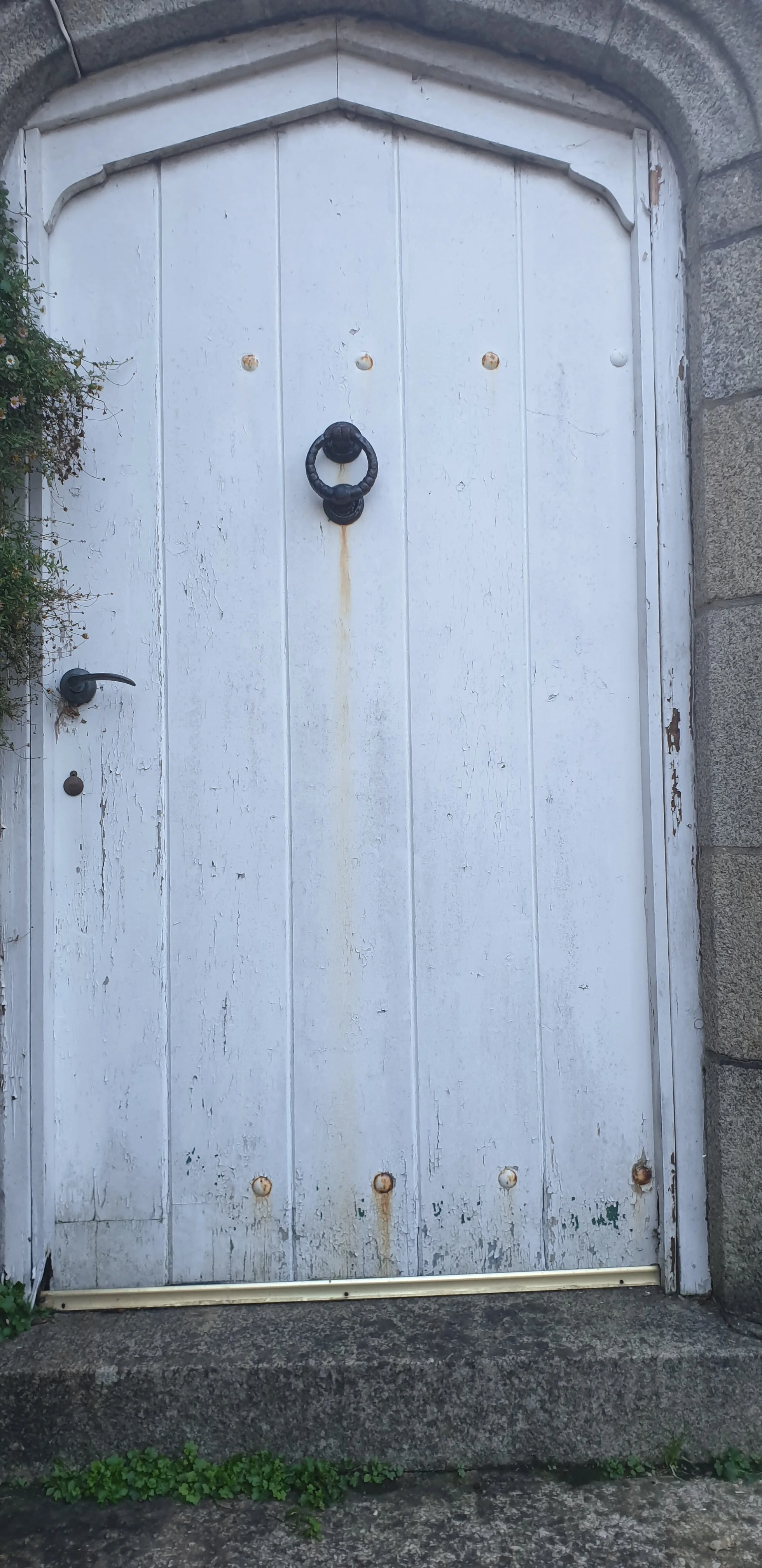 A white wooden door with a black door knocker and a black handle, framed by stone bricks with some chipped paint and rust spots, set on a concrete step with small green plants at the bottom.
