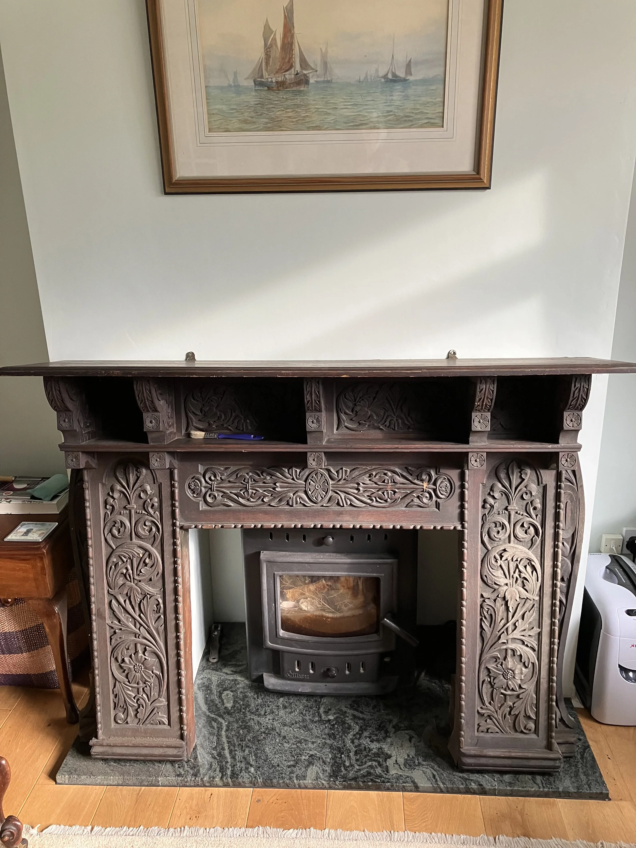 A fireplace surround with intricate wood carvings, with a modern wood-burning stove inside, situated on a green marble hearth under a framed nautical painting on a light-colored wall.