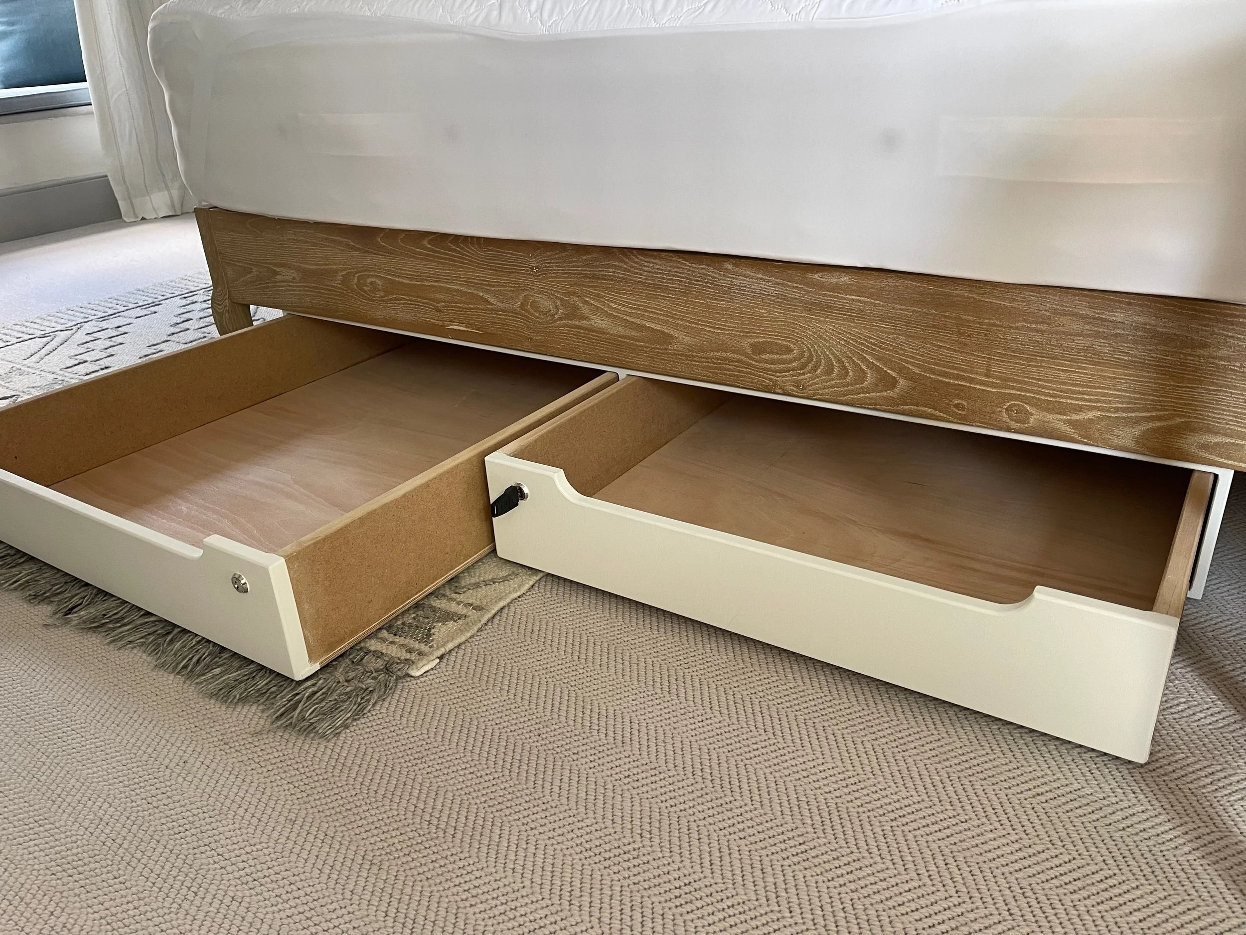 Open drawers under a bed, one white and one brown, revealing empty wooden compartments.