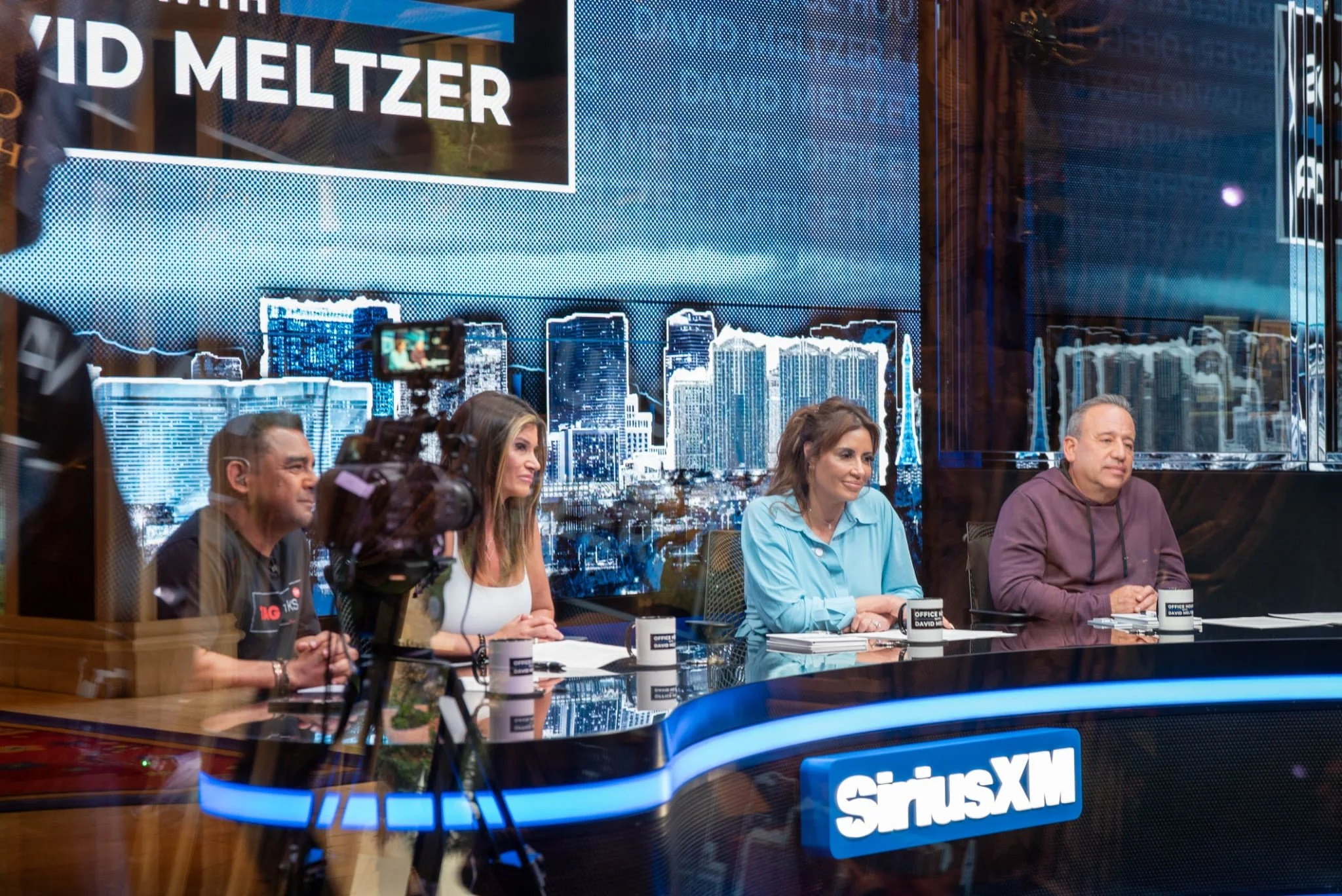People sitting at a SiriusXM radio station during a recording session, with a cityscape backdrop and a camera filming the event.