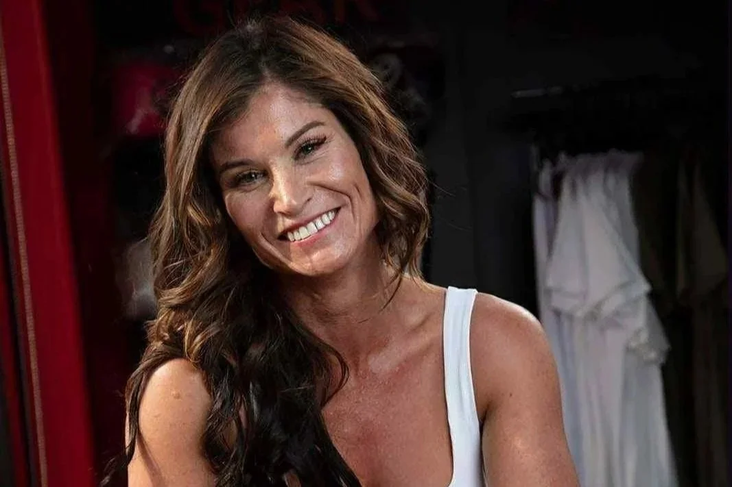 A woman with long wavy brown hair smiling, wearing a white tank top, in a room with hanging clothes in the background.