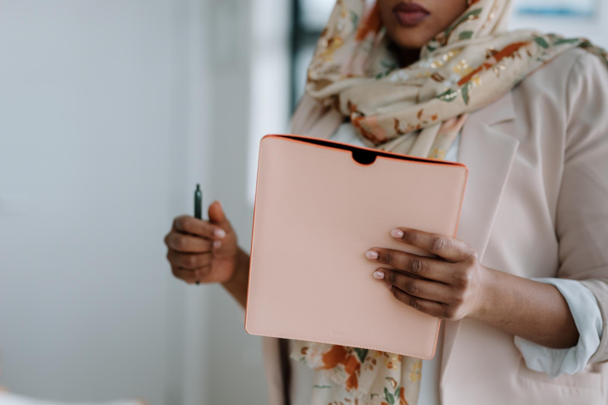 A woman holding a light pink notebook and a black pen, wearing a beige blazer and a floral scarf.