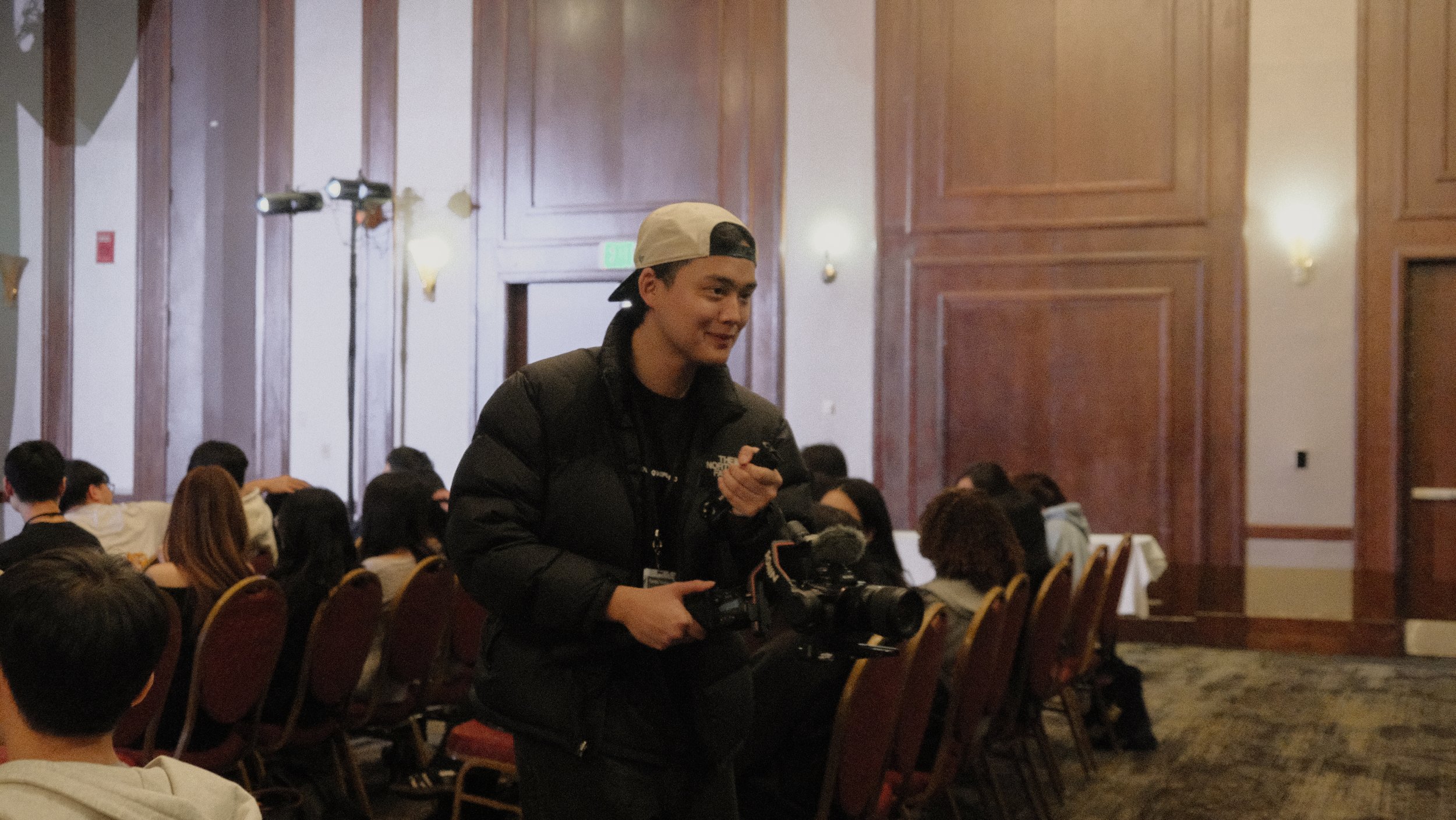 A young man holding a camera standing in a conference room filled with seated attendees.