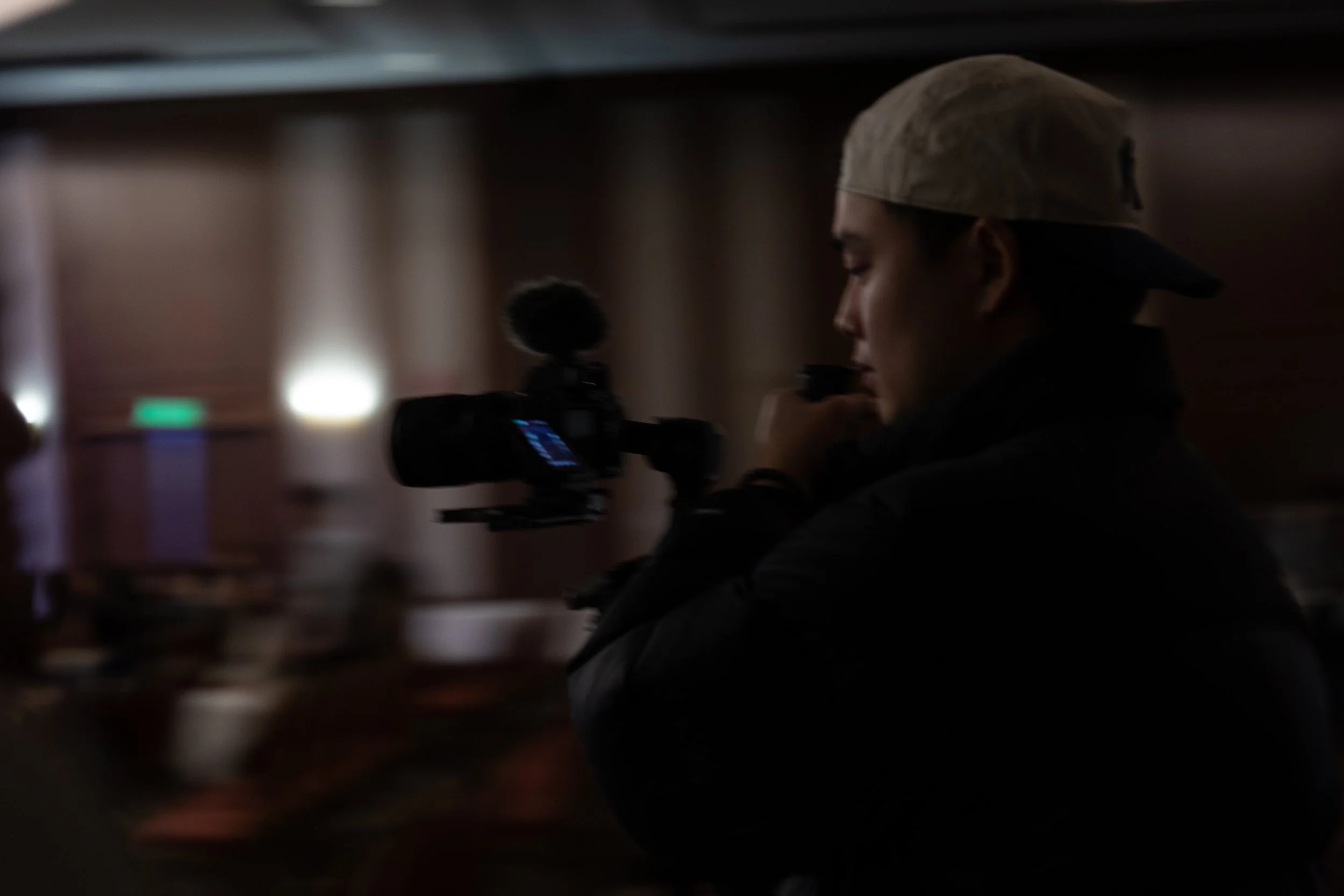 A person wearing a beige cap and black hoodie filming with a professional video camera in a dimly lit indoor setting.