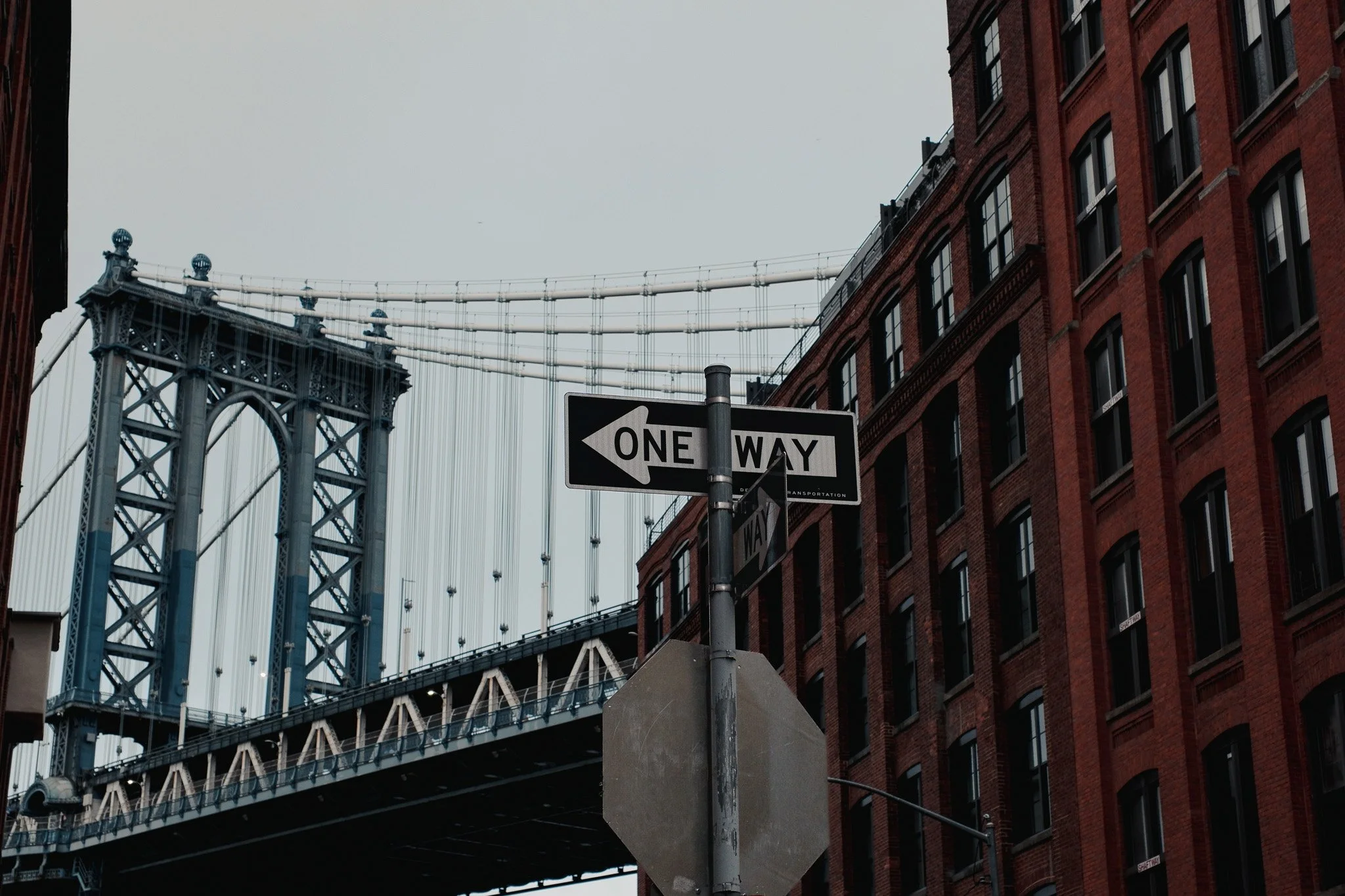 A one-way street sign in front of a brick building with the Brooklyn Bridge in the background.