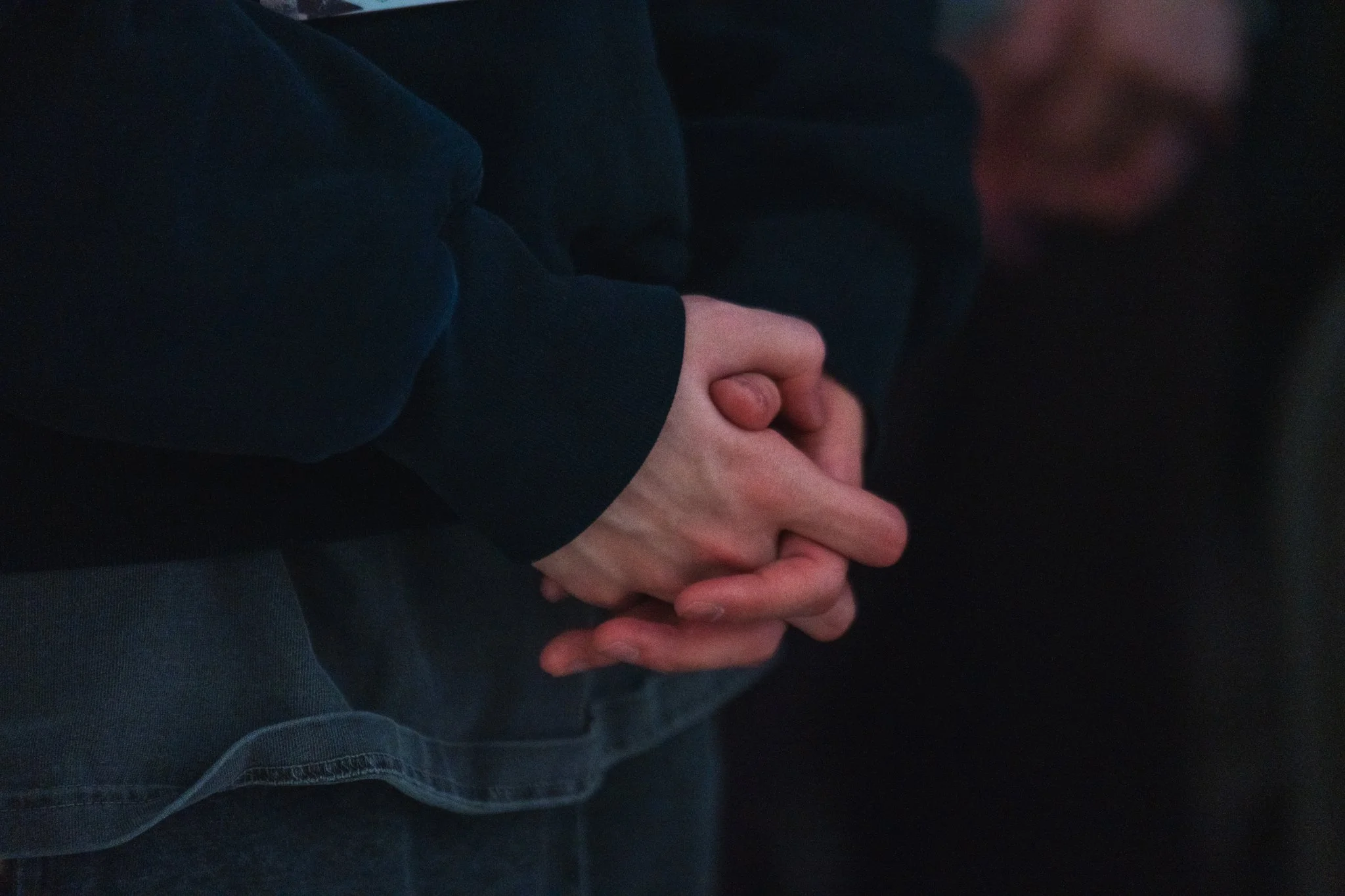 Close-up of person's hands clasped together in prayer, wearing a dark long-sleeve shirt, with a blurry face in the background.