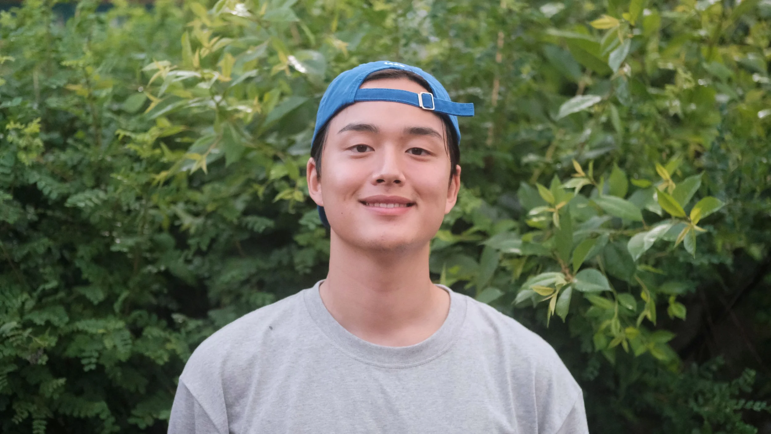 A young man with a slight smile, wearing a gray T-shirt and a blue cap backwards, standing in front of green foliage.