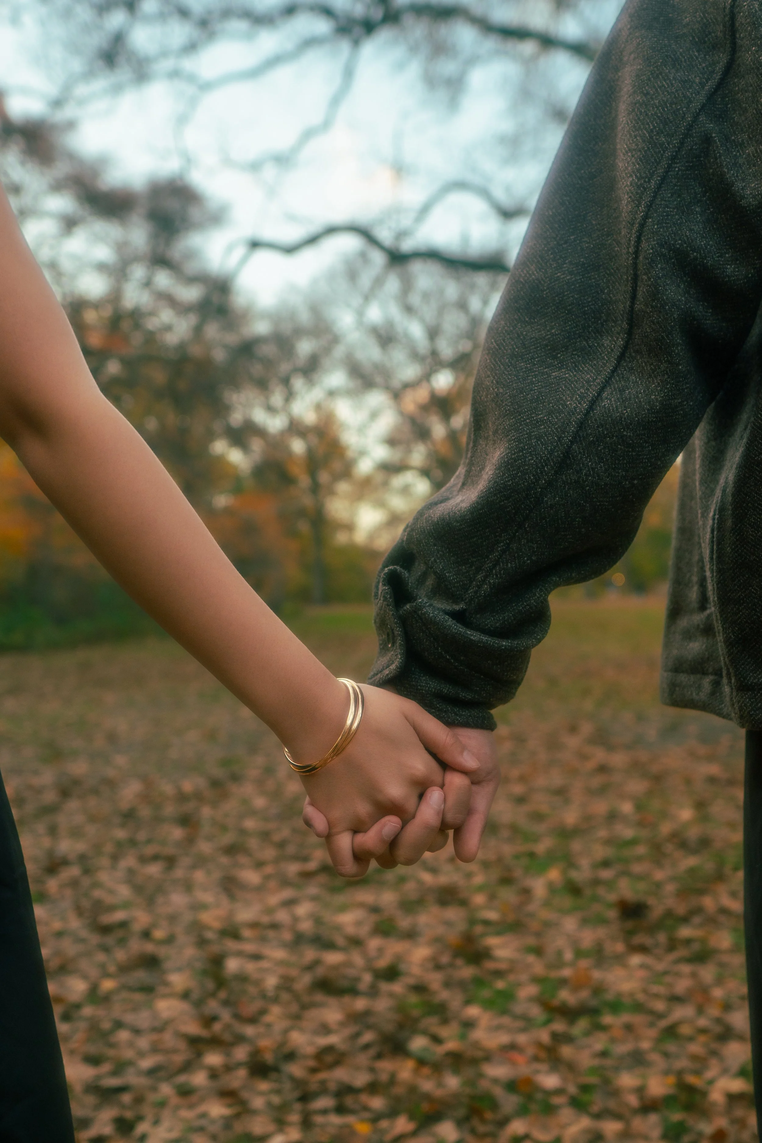 Close-up of a couple holding hands outdoors in a park during autumn, with trees and fallen leaves in the background.
