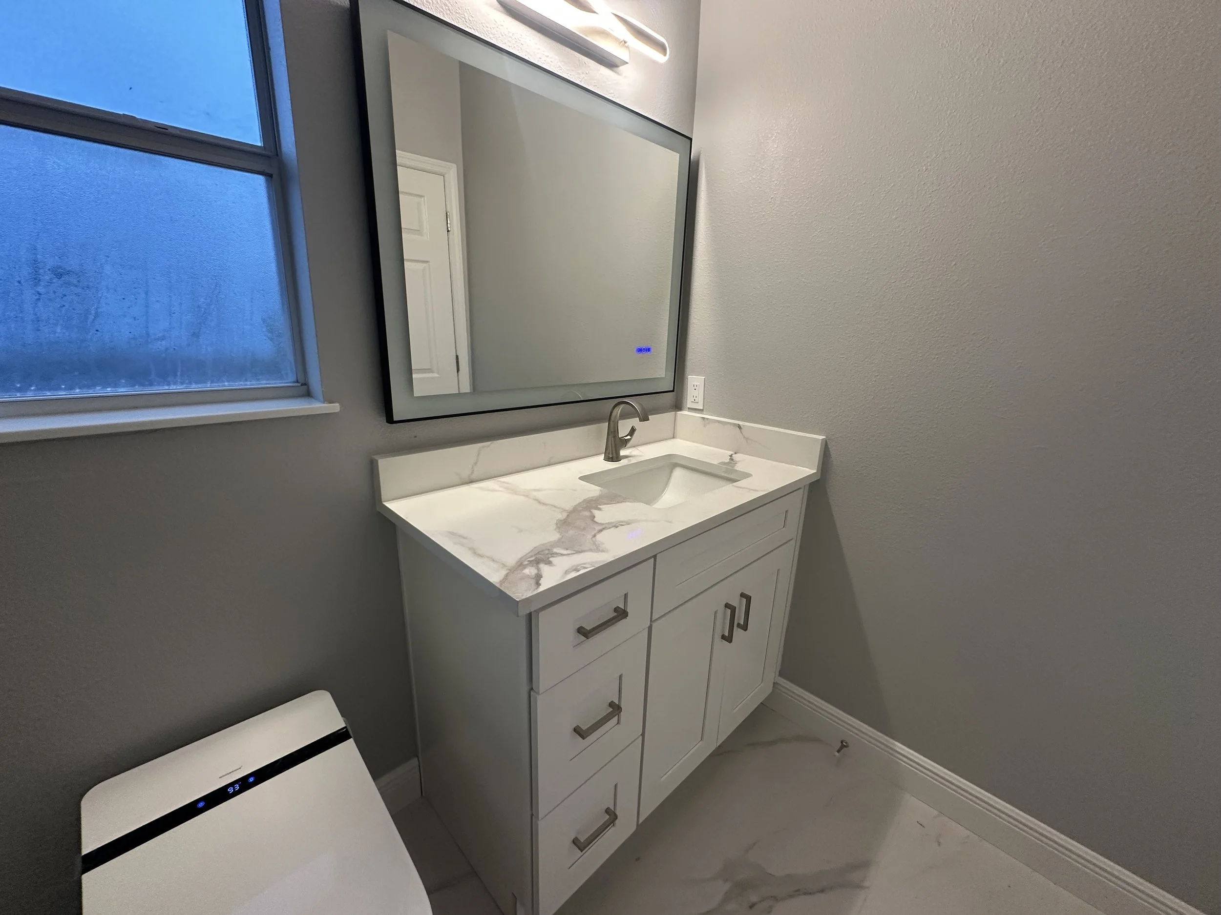 A modern bathroom vanity with a marble countertop, integrated sink, and brushed metal handles under a large mirror. To the left, a window with condensation and a closed door reflected in the mirror. Part of a white appliance with a digital display is visible in the lower left corner.