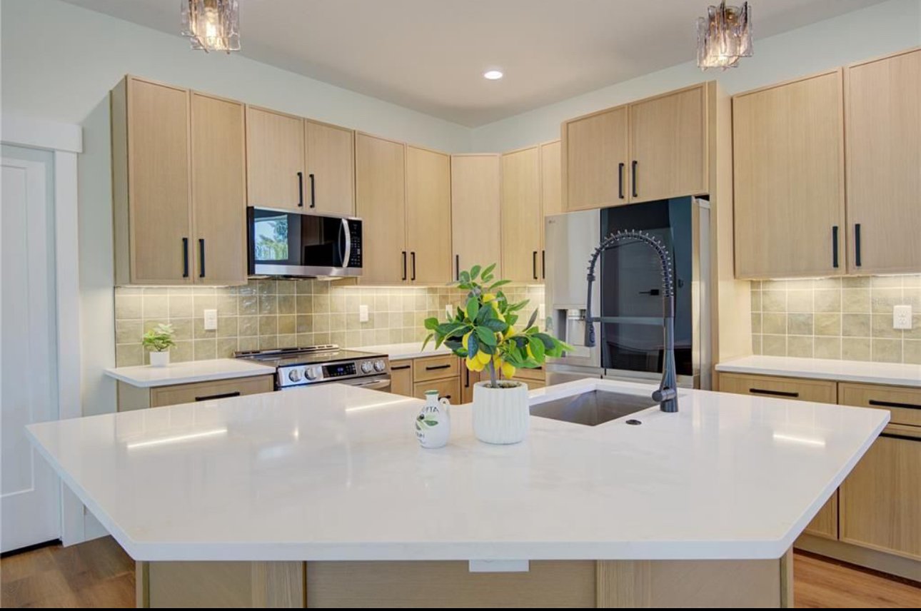 A modern kitchen with light wood cabinets, a white island with a sink and a black faucet, a microwave above a stove, and a plant on the island, with tiled backsplash and neutral-colored walls.
