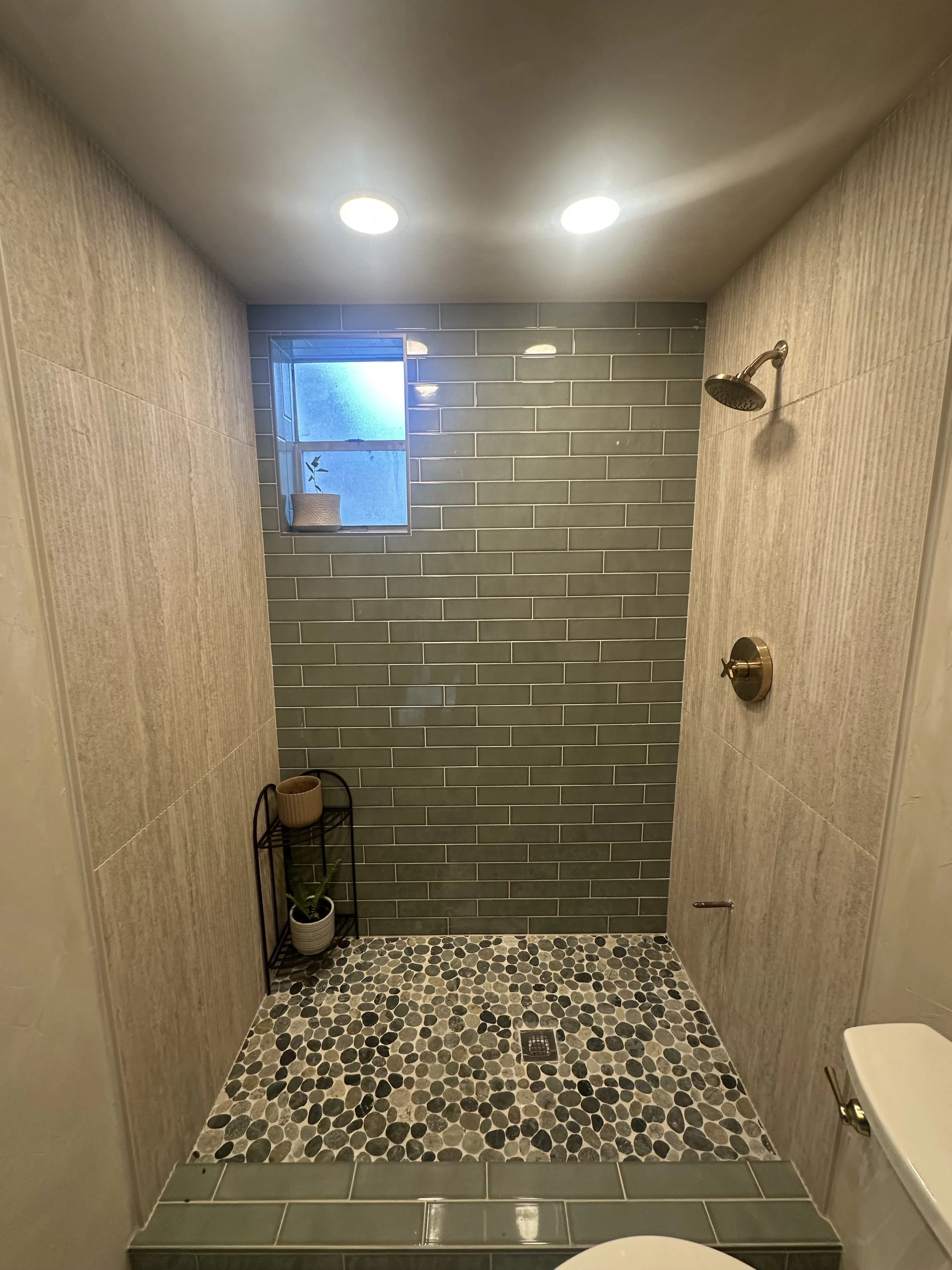 A walk-in shower with beige walls, a green tiled accent wall, small pebble-like tiles on the floor, a small window with a plant, and a wall-mounted showerhead.