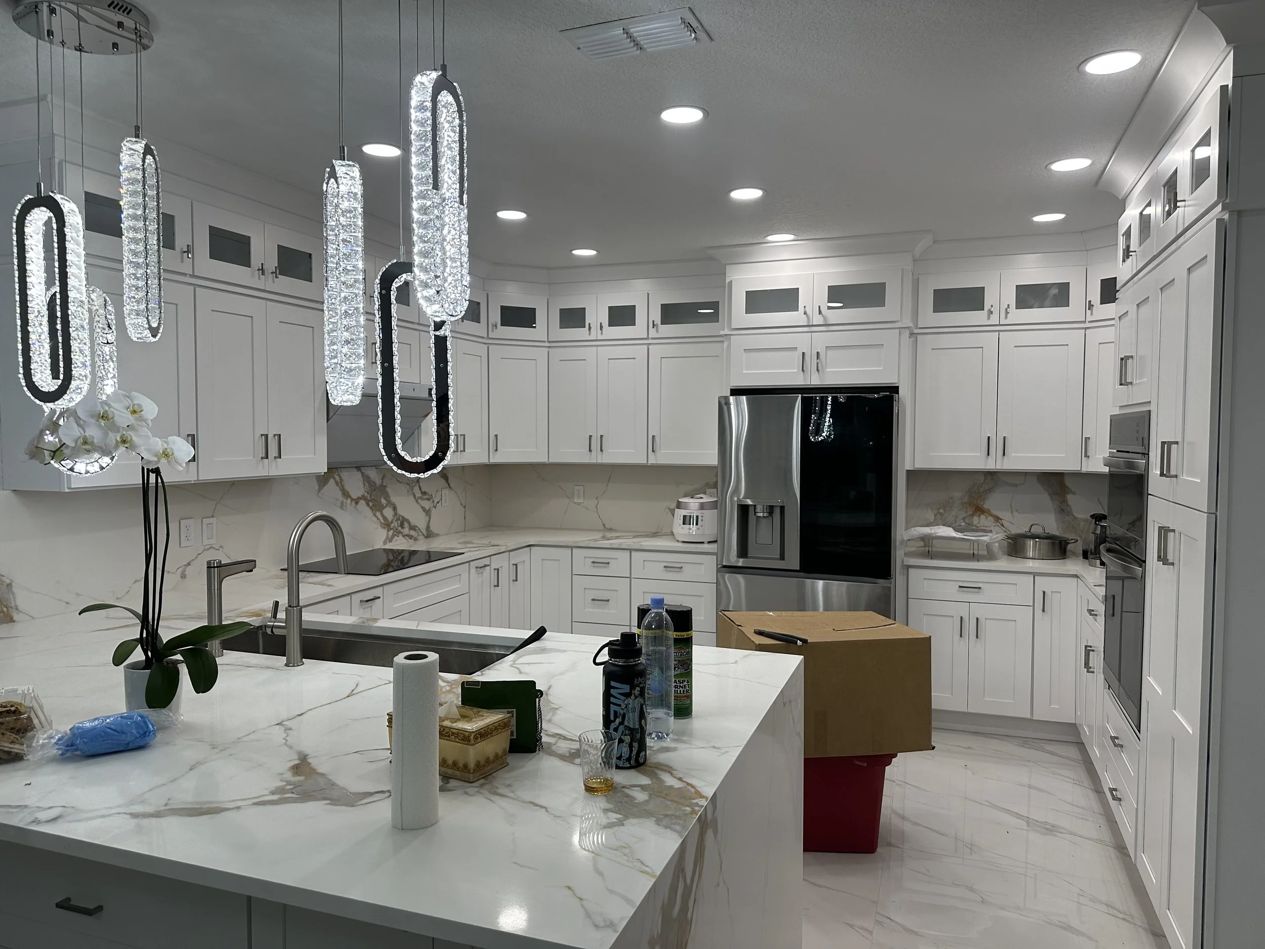 Modern kitchen with white cabinets, marble countertops, and stainless steel appliances. Hanging decorative lights over the island, various items on the island including bottles, tissue paper, and a box, with a flower in a pot on the counter.