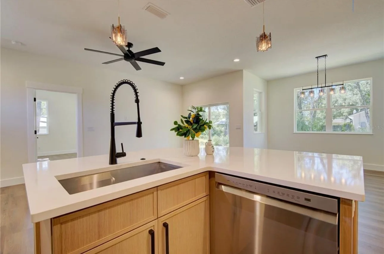 Modern kitchen with a white countertop island, black faucet, wooden cabinetry, stainless steel dishwasher, decorative plant, and large windows.