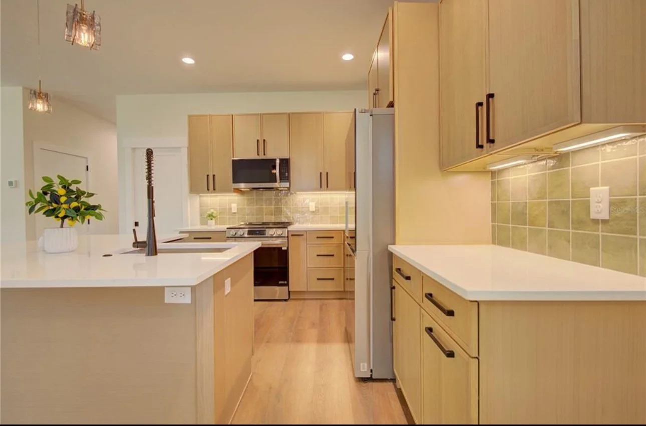 Kitchen with wooden cabinets, white countertops, a gray refrigerator, oven, microwave, and a small plant on the counter. There's a backsplash with beige tiles and under-cabinet lighting.