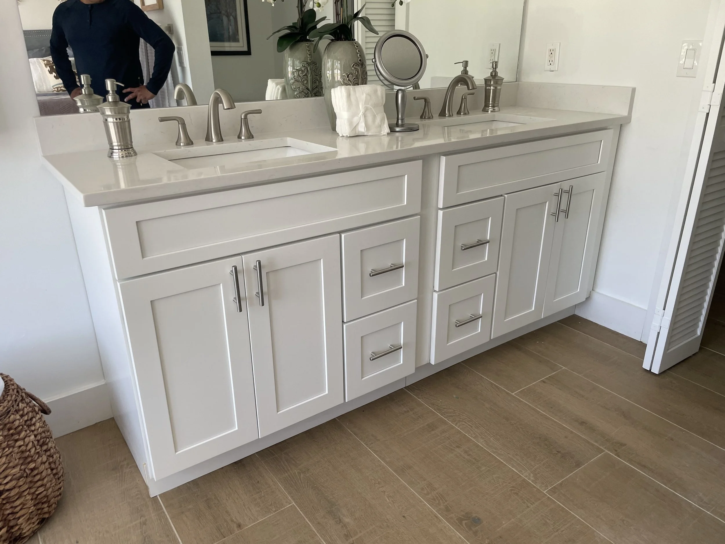 White bathroom vanity with two sinks, silver faucets, and a large mirror. There are decorative vases and a mirror on the counter, with a person standing in reflection visible in the mirror. The vanity has cabinets and drawers with silver handles, and the floor is wooden.