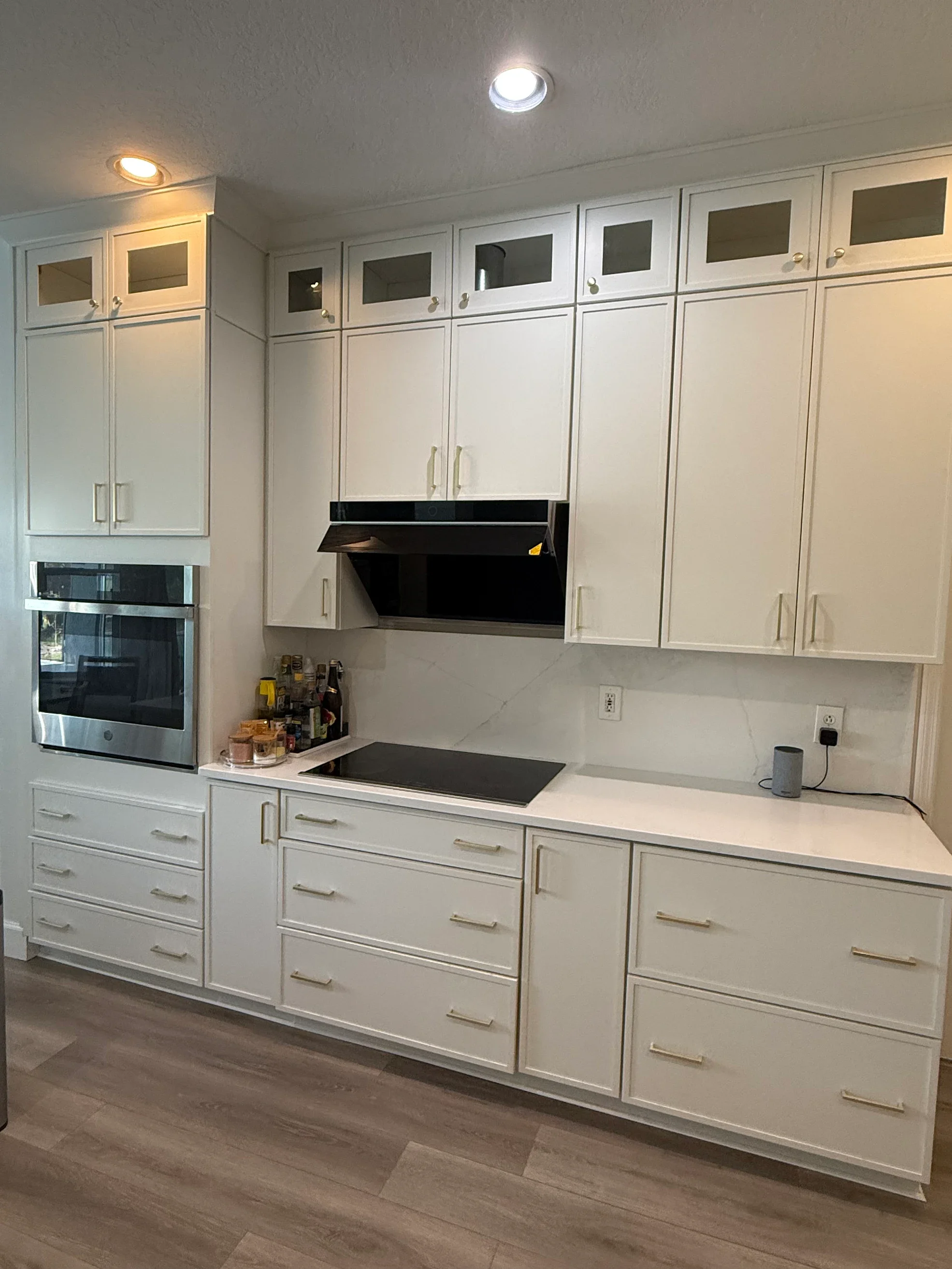 White kitchen cabinets with gold handles, a black stove and hood, a built-in oven, and a countertop with bottles, jars, and a speaker. Finished with wood-look flooring.