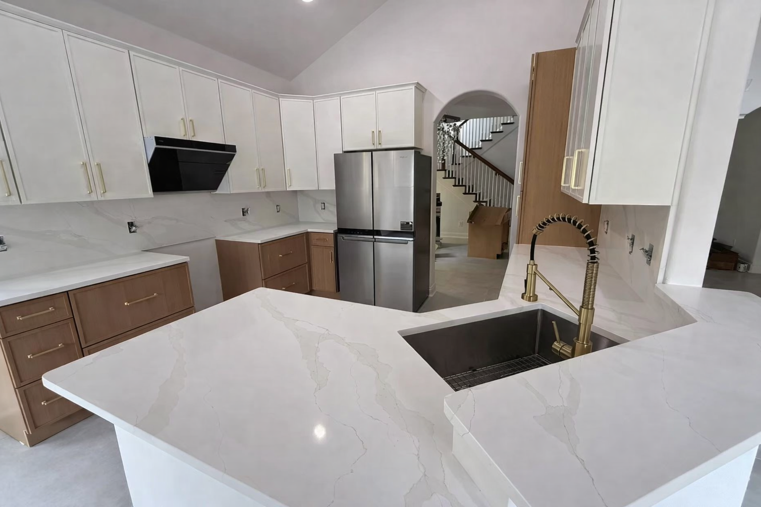 Modern kitchen with white and wood cabinets, stainless steel refrigerator, marble countertops, and a gold faucet over a black sink, with a staircase visible in the background.
