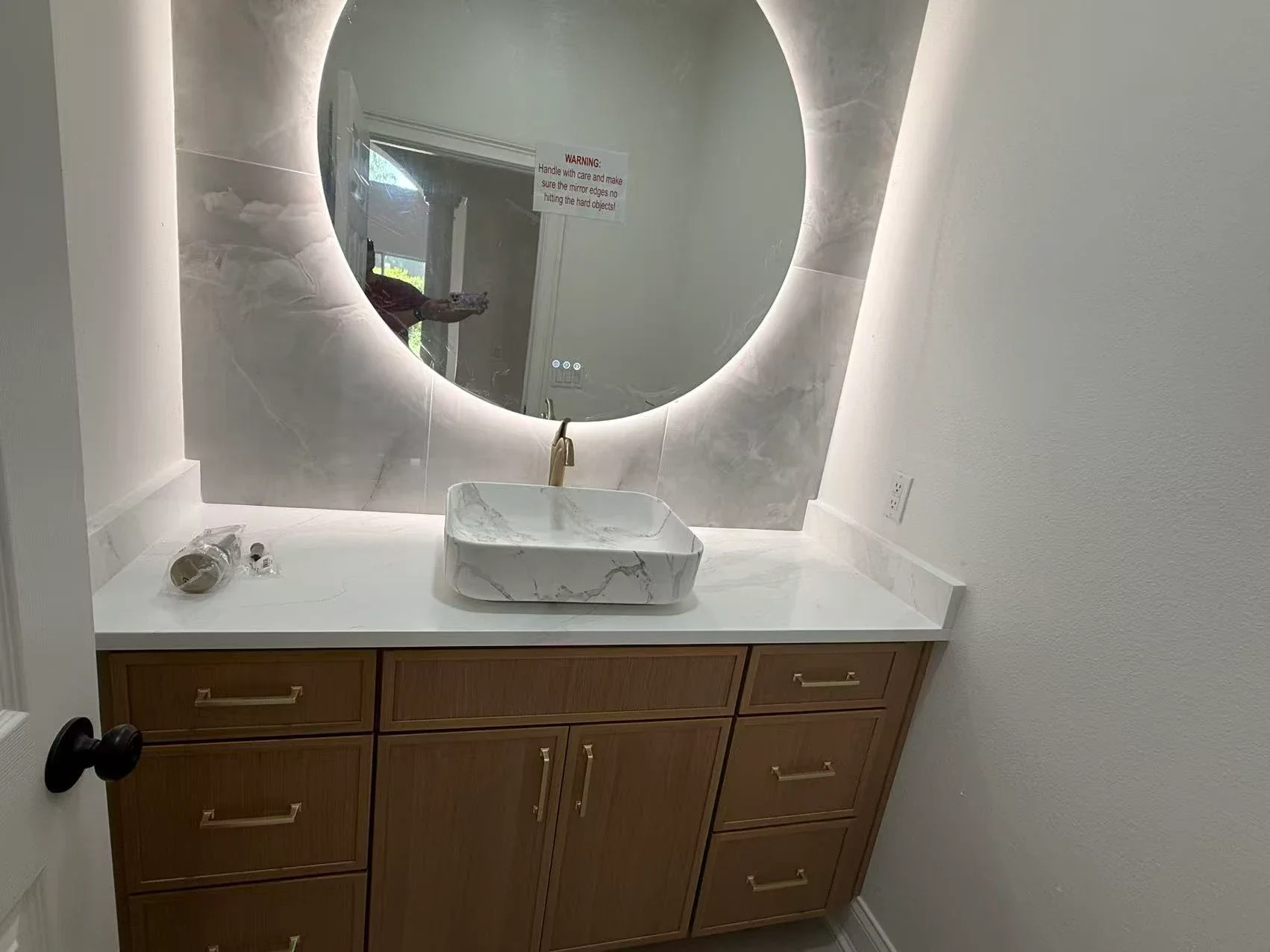 Bathroom vanity with a wooden cabinet, a white marble countertop, a marble sink, and a round backlit mirror. There is a faucet and some items on the countertop, and a warning sign on the mirror.